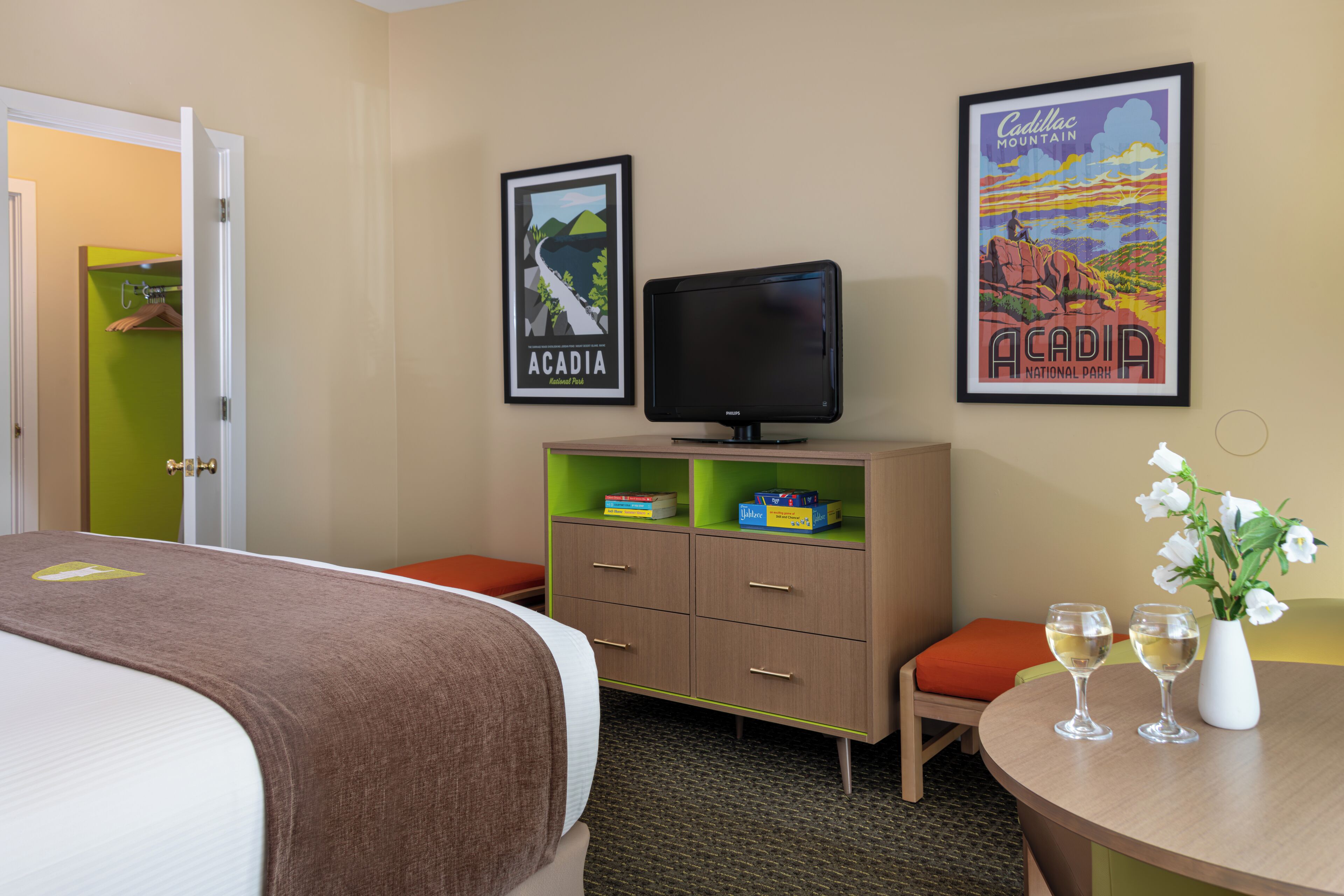 Een hotelkamer met een bed, een bureau met twee glazen wijn, een televisie, een kast en twee ingelijste posters aan de muur.