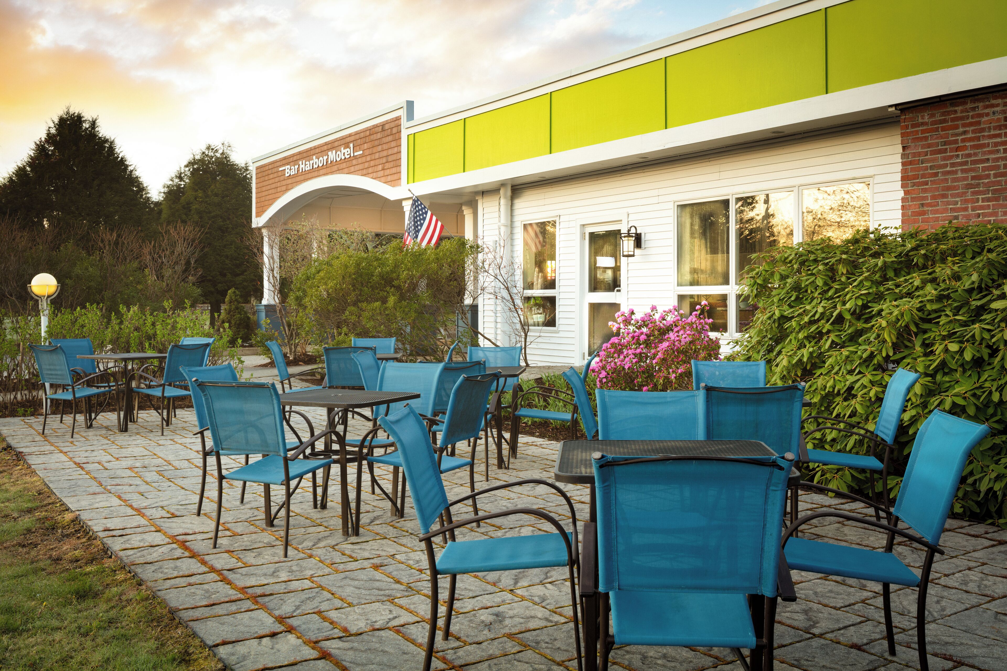 Un patio al aire libre con sillas y mesas azules, una bandera estadounidense y un edificio que dice "Bar Harbor Motel".