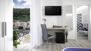A modern hotel room with a balcony, a desk with a chair, a TV mounted on the wall, and a view of the city and hills.