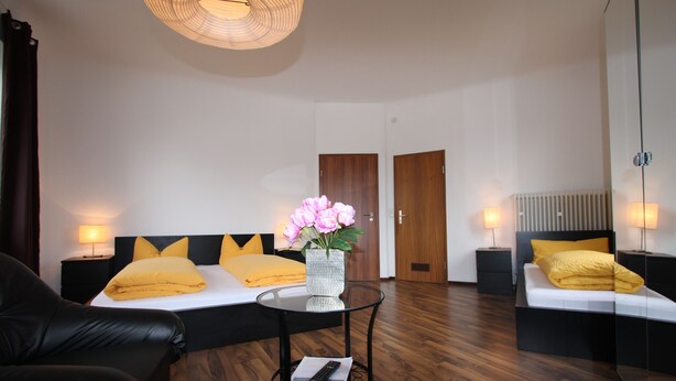 A modern hotel room with a bed, two bedside tables, a small round table, a sofa, and a decorative vase with flowers. - Stadthotel Kleiner Berg (Friedrichshafen)