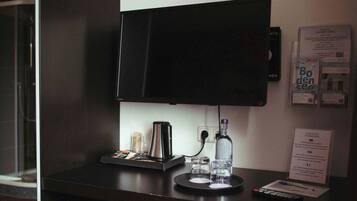 Ein Hotelzimmer mit einem an der Wand befestigten Flachbildfernseher, einer Kaffeemaschine, einer Wasserflasche und einer Fernbedienung auf dem Schreibtisch.