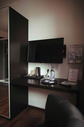 A hotel room with a flat-screen TV mounted on the wall, a coffee maker, a bottle of water, and a remote control on the desk.