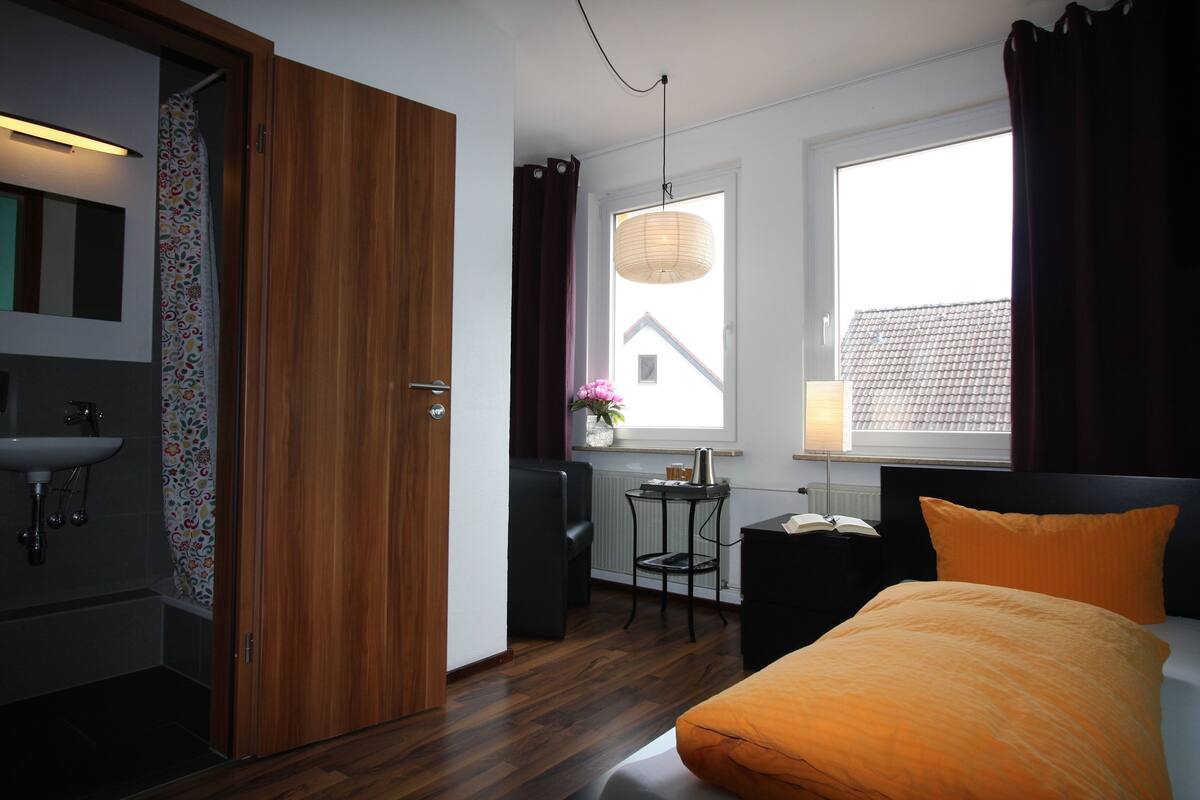 A hotel room with a wooden door, a small table with a lamp, a bed with an orange pillow, and a view of the outside through a window.