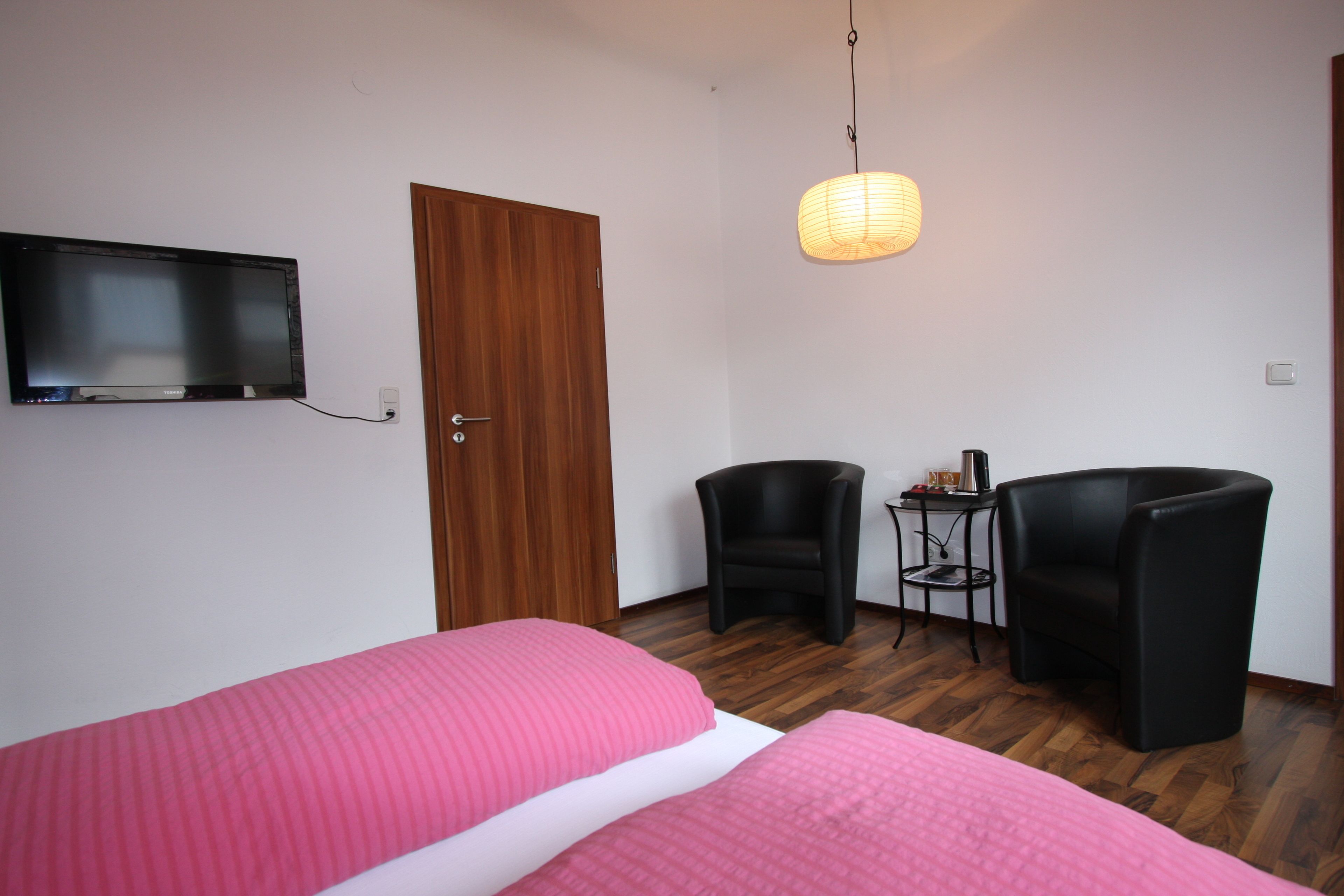Habitación de hotel con dos camas, televisor, puerta de madera, dos sillas negras, una mesita con lámpara y una lámpara colgante.