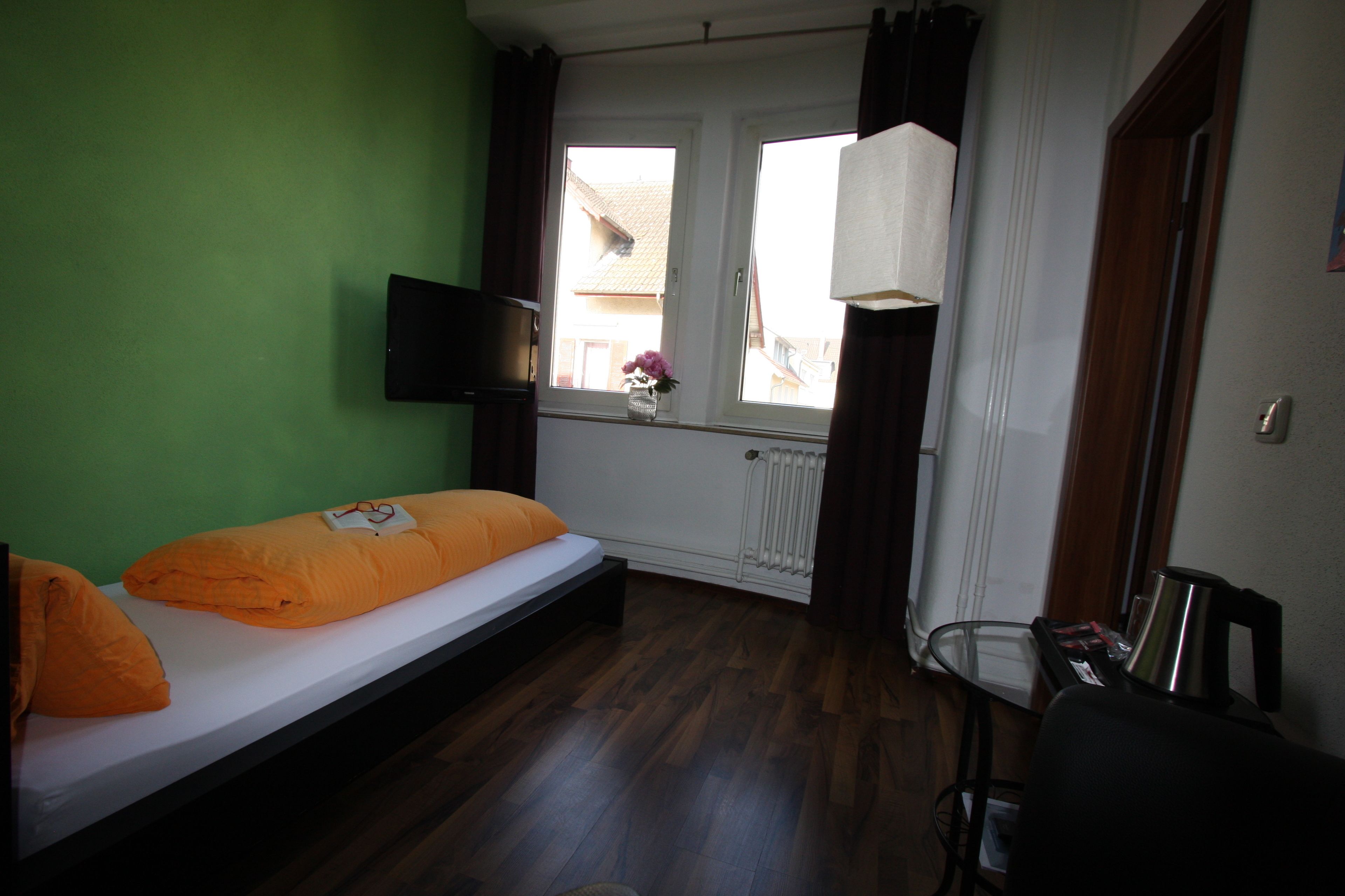 A room with a single bed, a small table with a kettle, a chair, and a window with curtains.