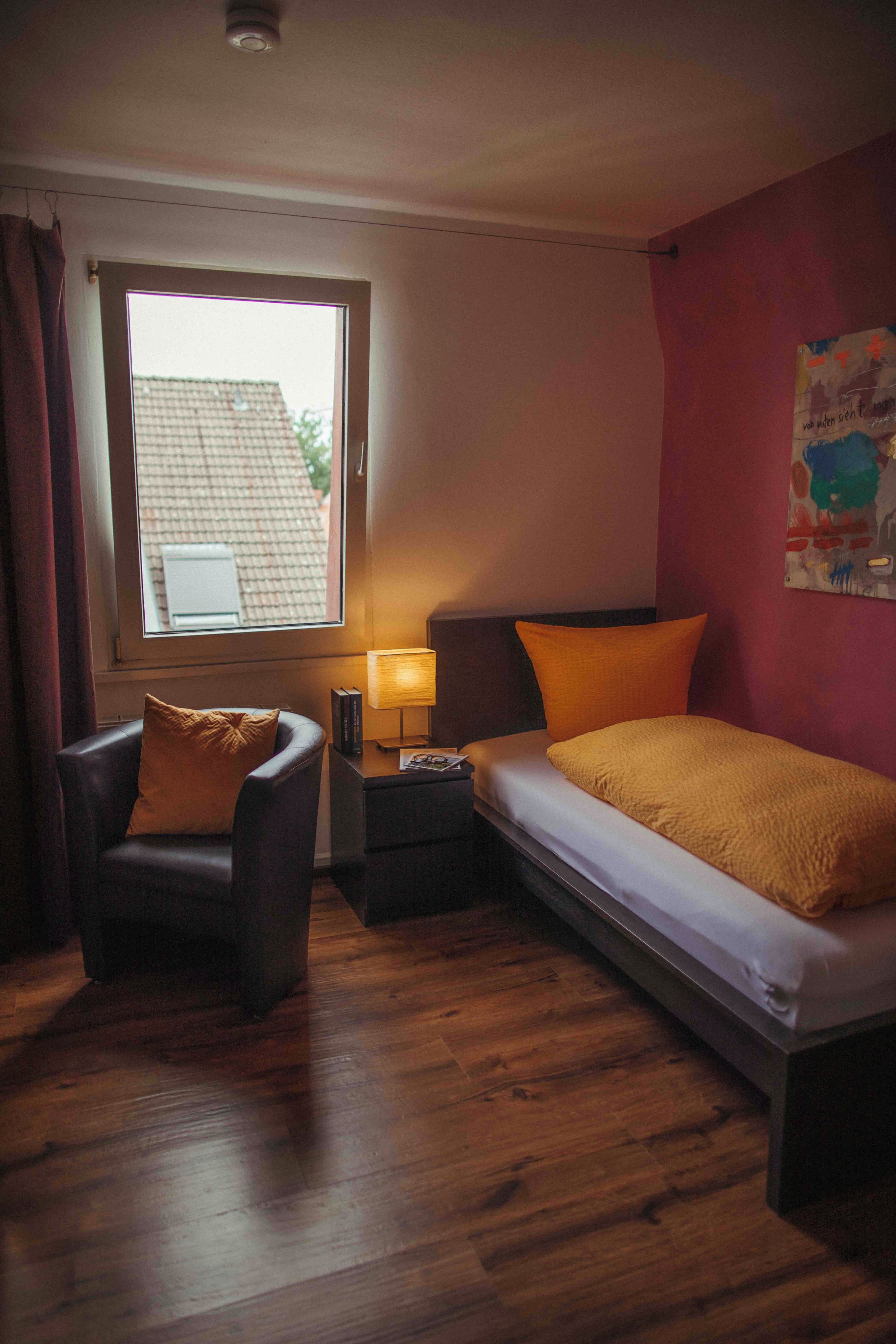 Une chambre d’hôtel avec un lit, une chaise, une table de chevet, une lampe et une fenêtre.
