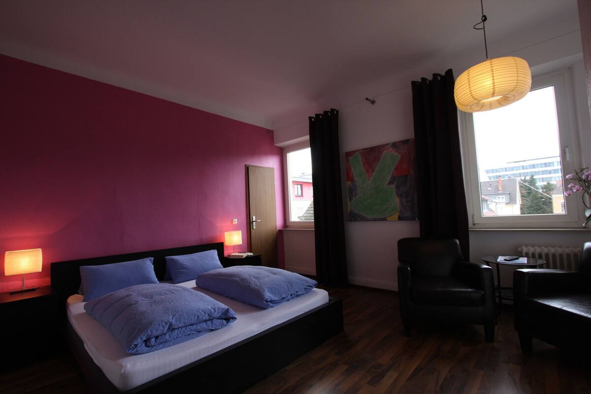 A bedroom with a bed, two bedside tables, a chair, a small table, a window with curtains, and a hanging light fixture.