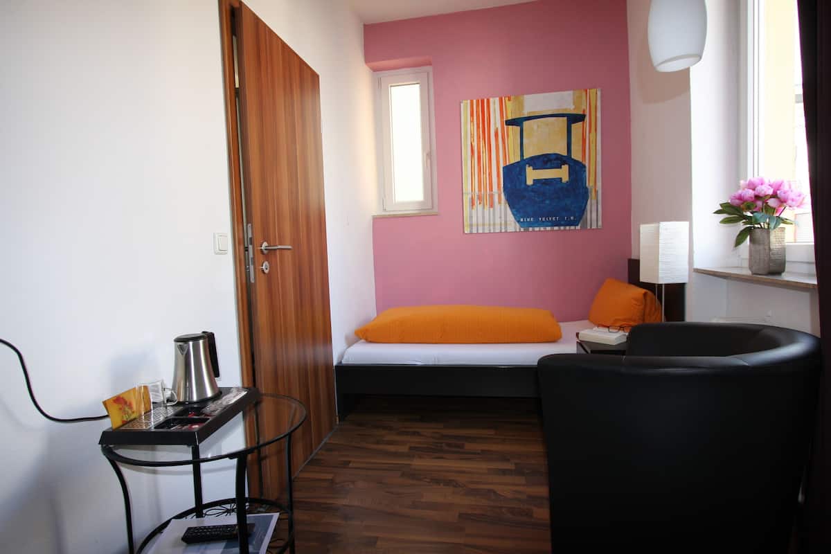 A small room with a bed, a chair, a table with a kettle, a window, and a painting on the wall.
