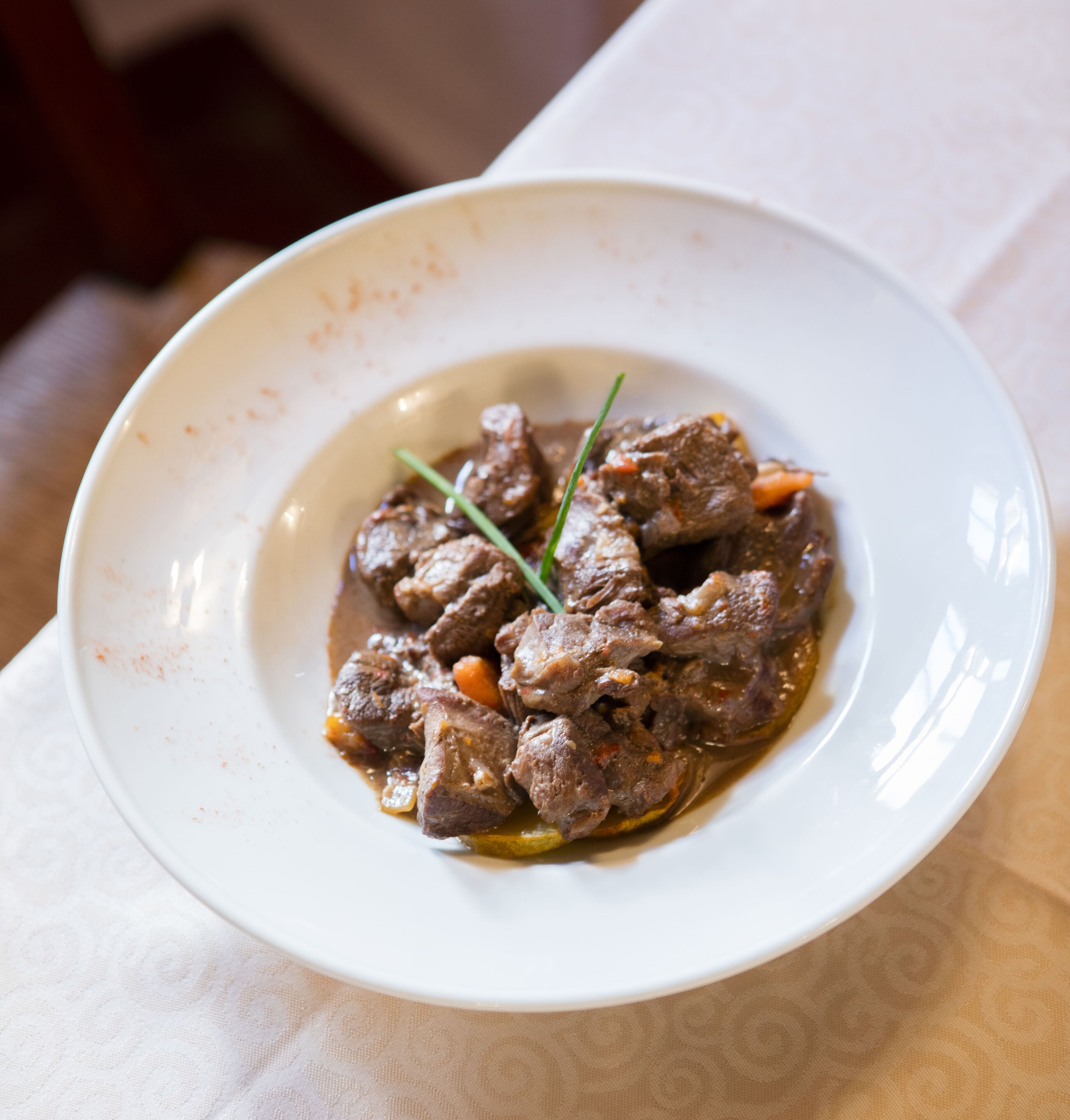A white bowl containing a beef dish with vegetables and a garnish, placed on a table with a patterned tablecloth.
