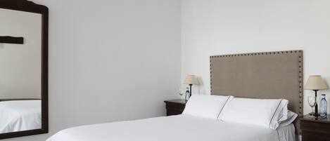 Una cama bien hecha con sábanas blancas, un espejo grande y dos mesitas de noche con lámparas y botellas.