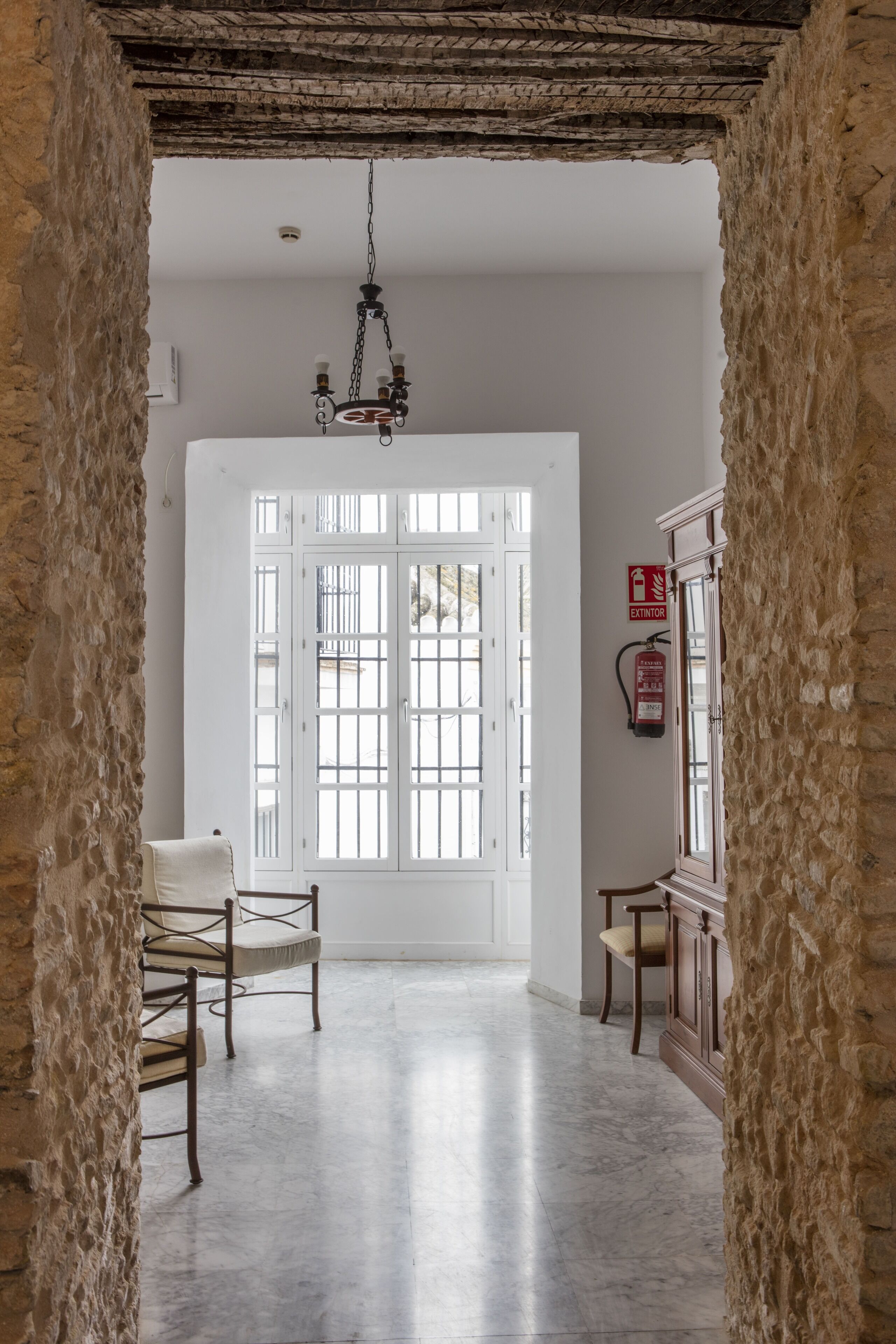 Un pasillo con paredes de piedra, una puerta blanca con paneles de vidrio y dos sillas de madera.