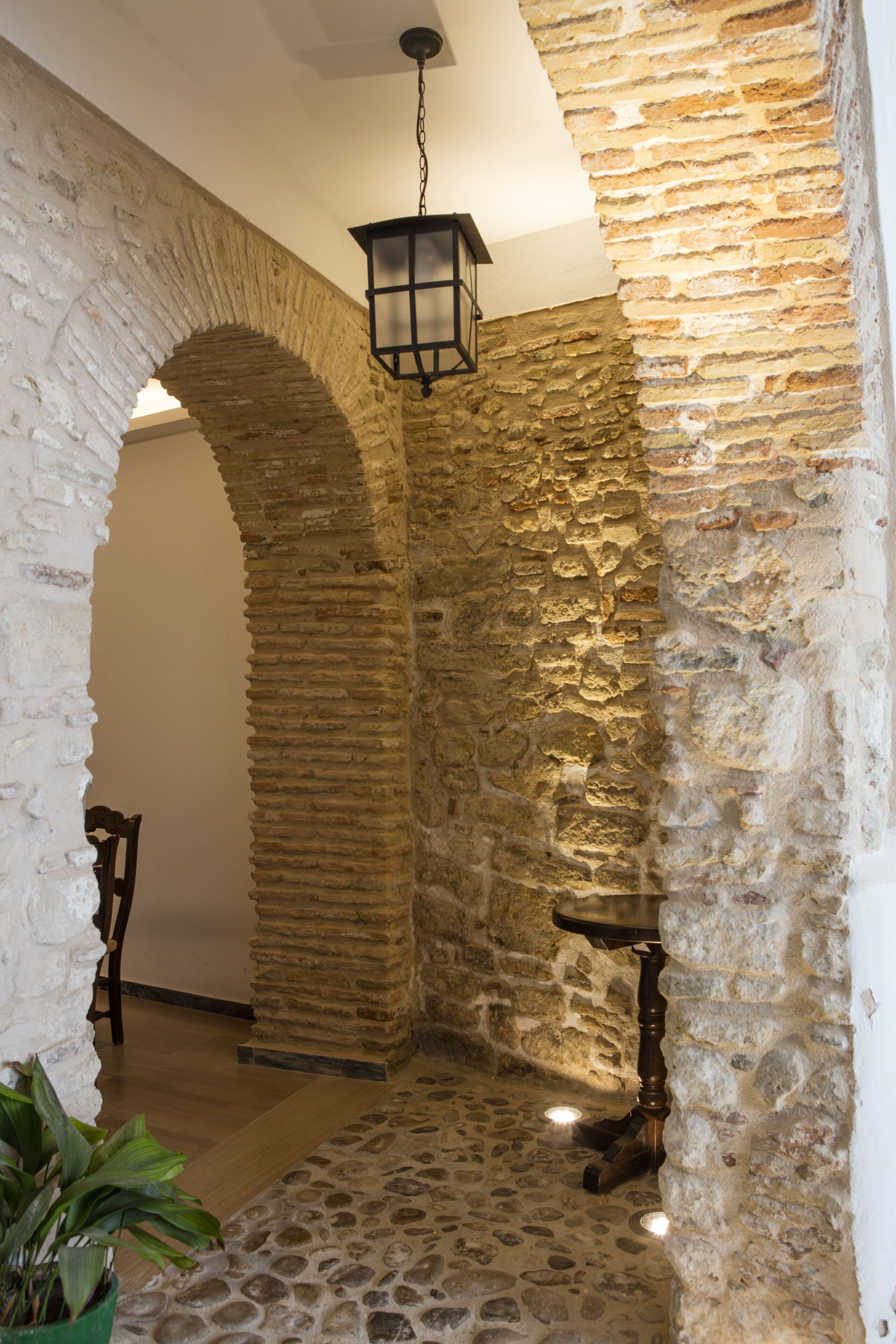 Un pasillo de paredes de piedra con un farol colgante, una silla de madera y una planta en maceta.