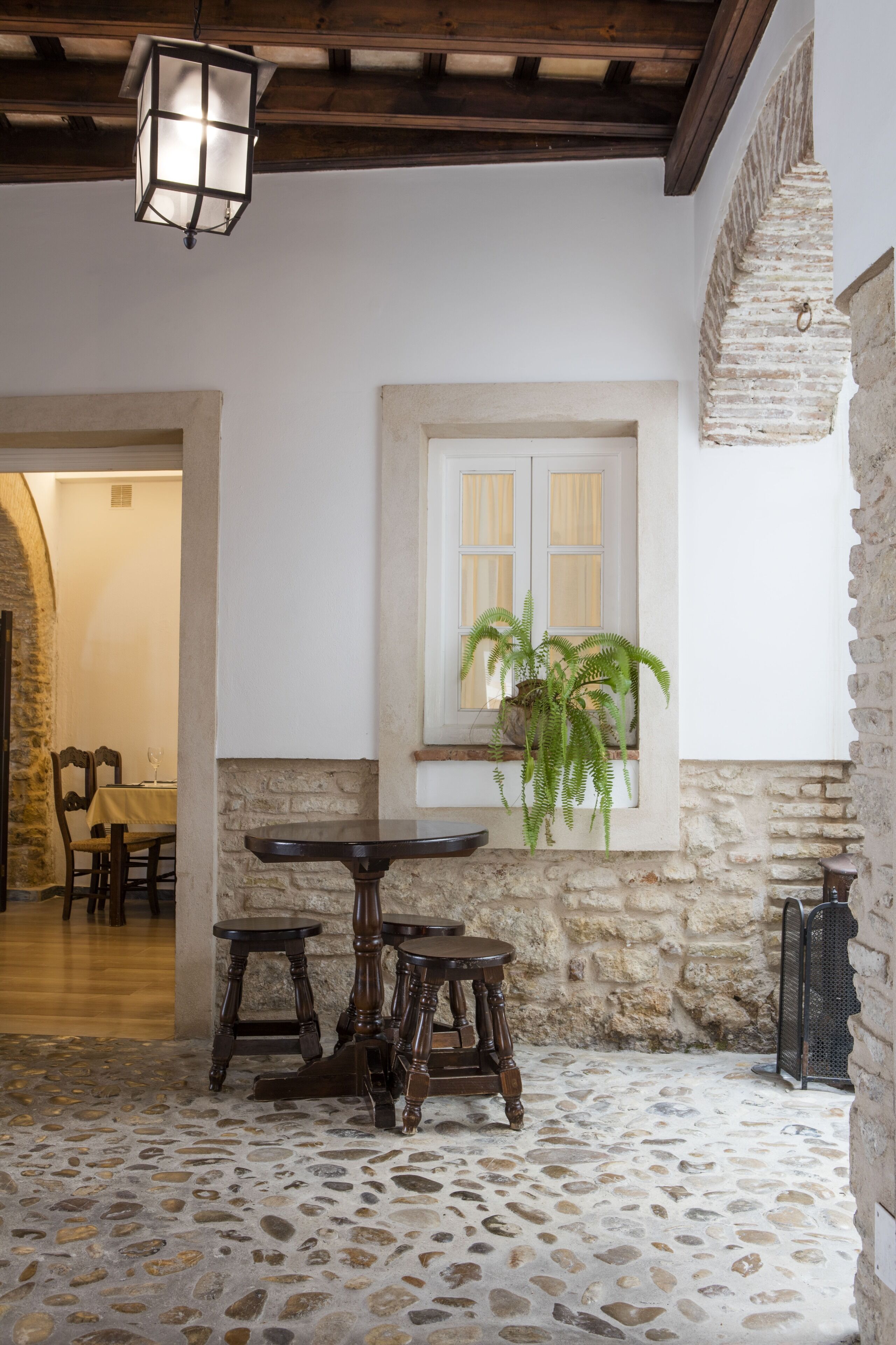 Un patio con suelo de piedra, una mesa y sillas de madera, un farol colgante y una ventana con una planta.