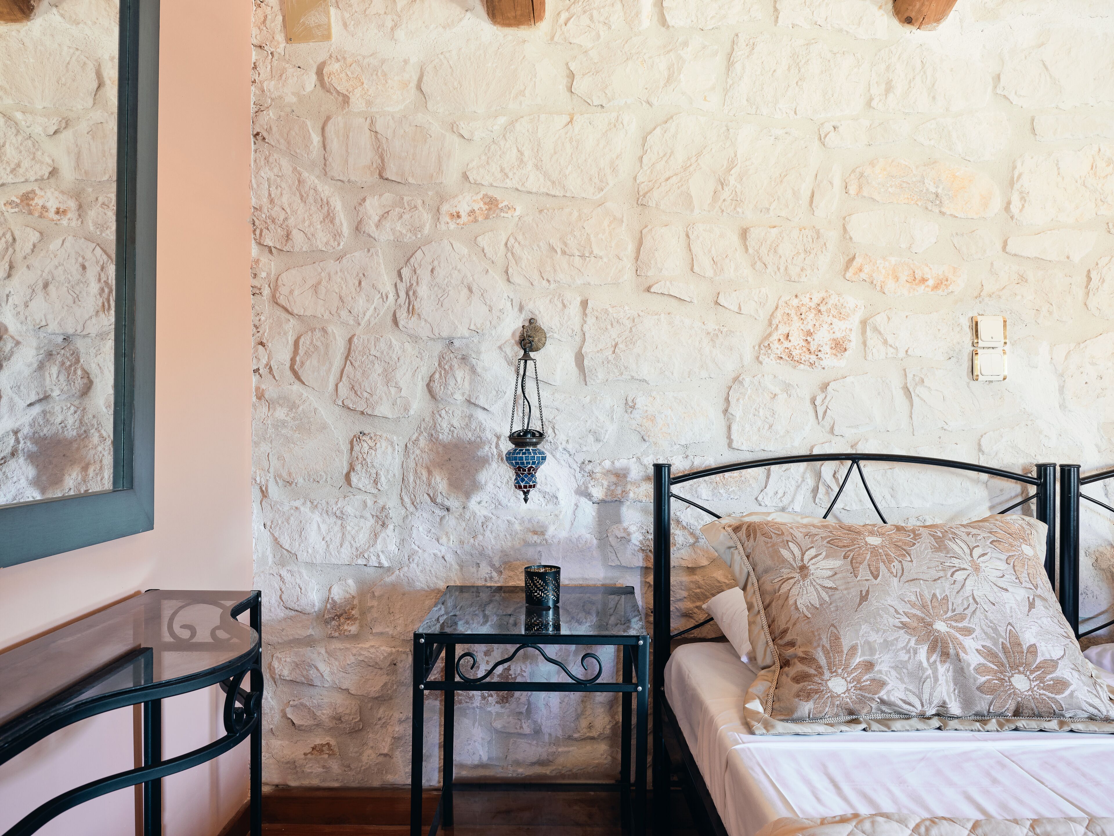 A room with a stone wall, a bed with floral pillows, a small table with a lamp, and a mirror on the wall.
