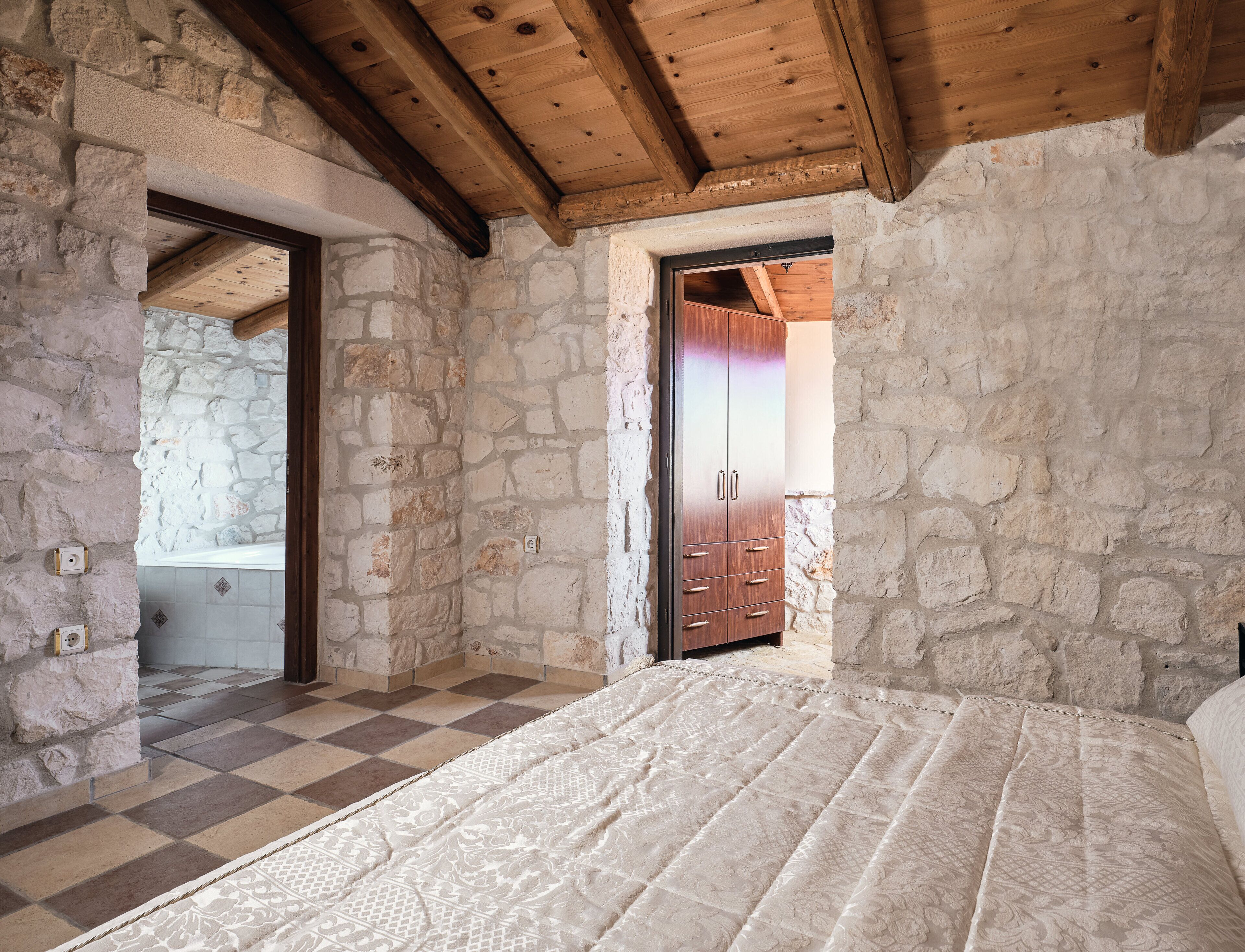 A bedroom with a checkered floor, stone walls, wooden beams, and a wardrobe.