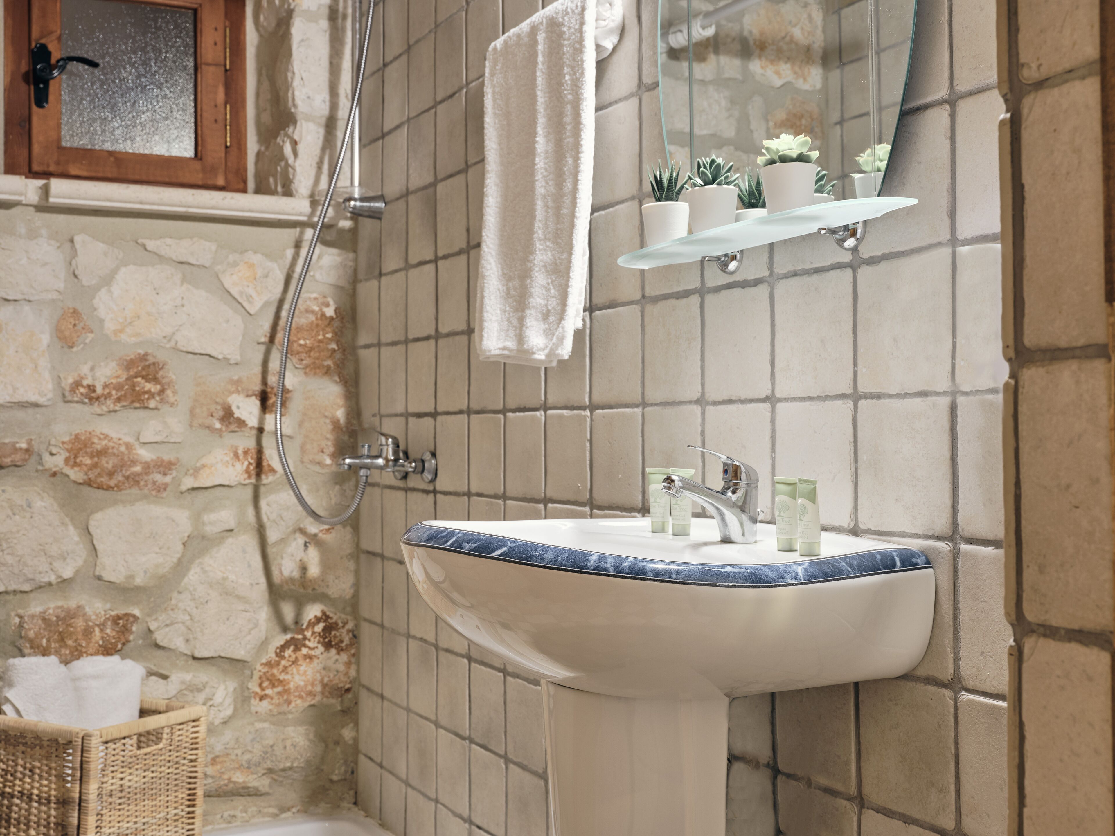 A bathroom with a stone wall, a white sink with blue trim, a shower, and a shelf with toiletries.