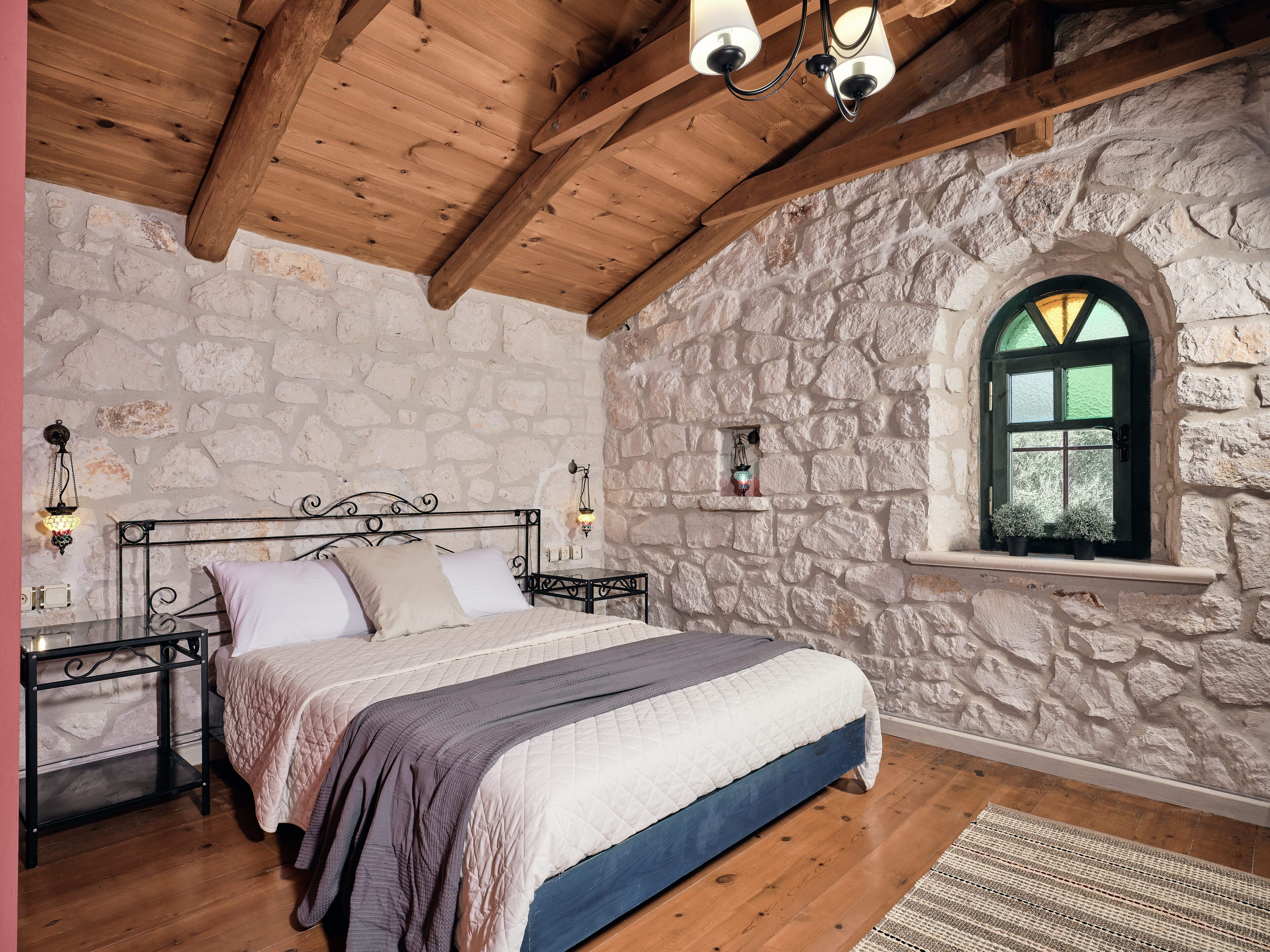 A bedroom with a stone wall, wooden ceiling beams, a bed with a quilted cover, a nightstand, and a window with a view.