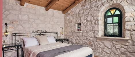 A bedroom with a stone wall, wooden ceiling beams, a bed with a quilted cover, a nightstand, and a window with a view.