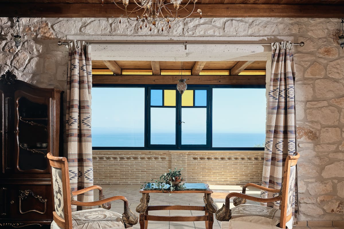 A room with a stone wall, a wooden ceiling, and a large window with a view of the sea. There are two wooden chairs and a small table with a plant.