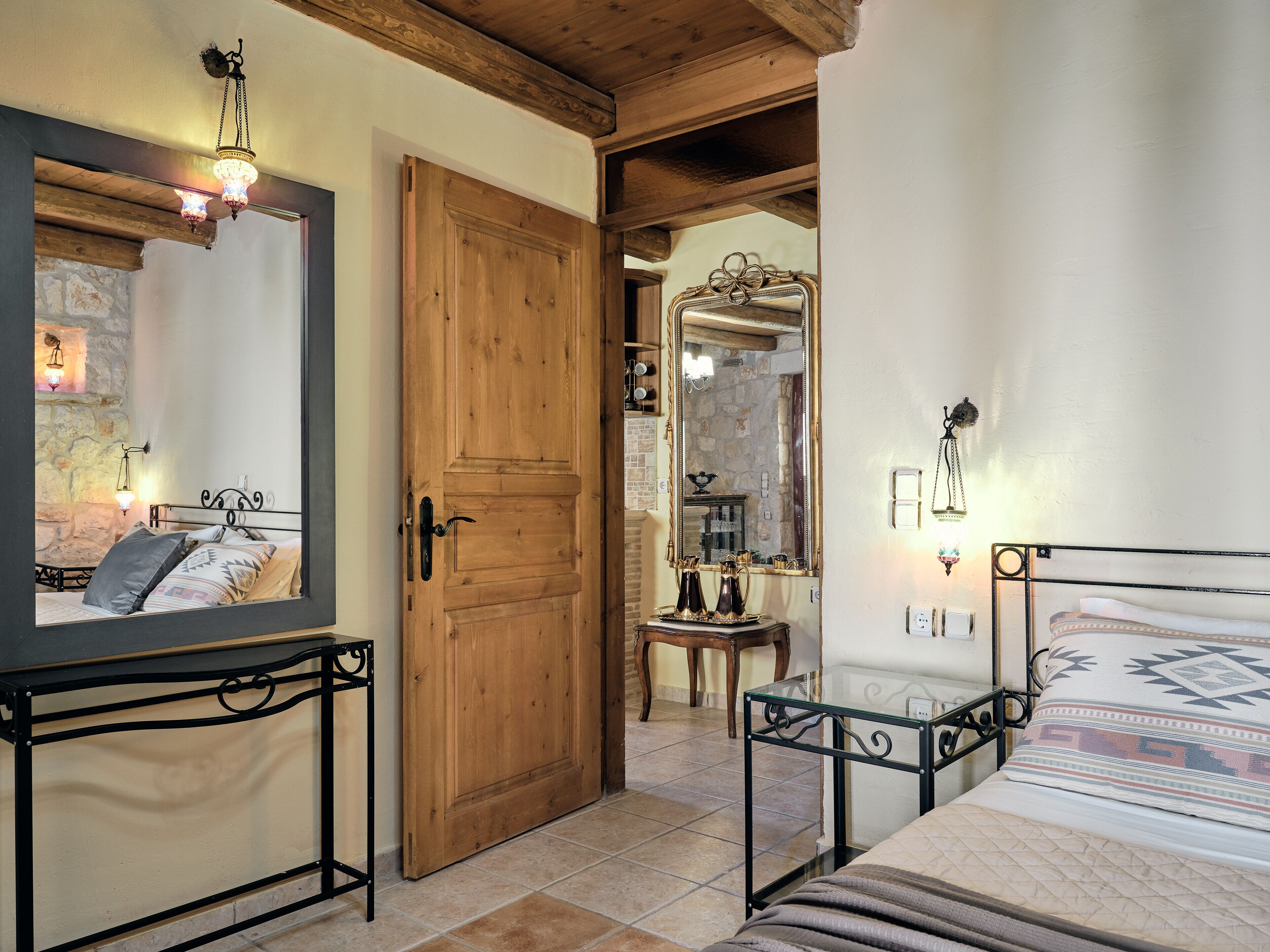 Une chambre avec une porte en bois, un lit recouvert d’une couvre-lit à motifs, une table de chevet, un miroir et une petite table ornée d’objets décoratifs.