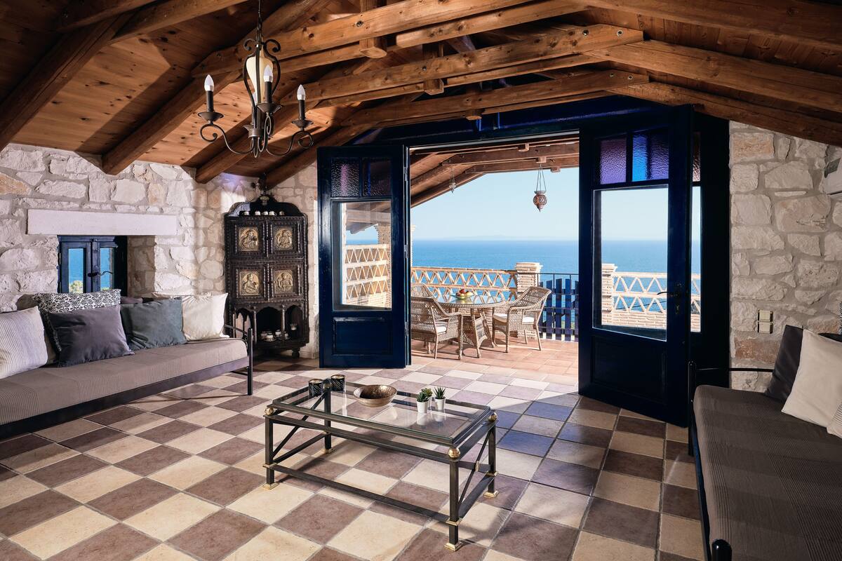A cozy room with a checkered floor, a wooden ceiling, a stone wall, a sofa, a table, and a view of the sea.