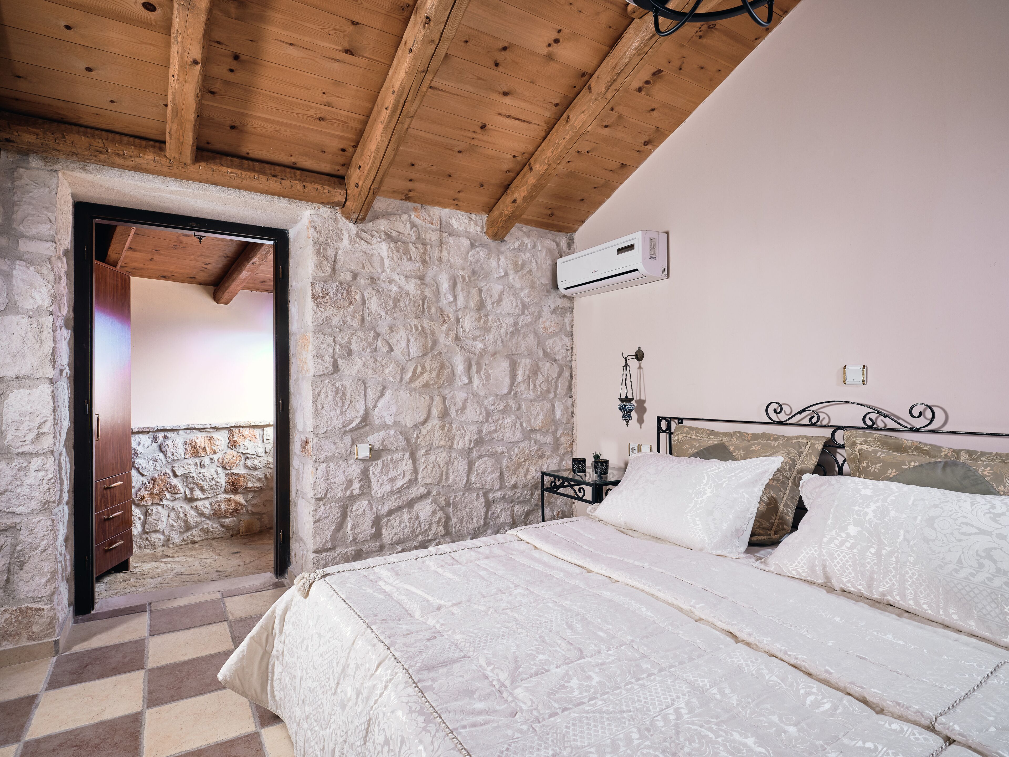 A bedroom with a stone wall, wooden ceiling beams, a bed with patterned bedding, a bedside table with a lamp, and an air conditioning unit.
