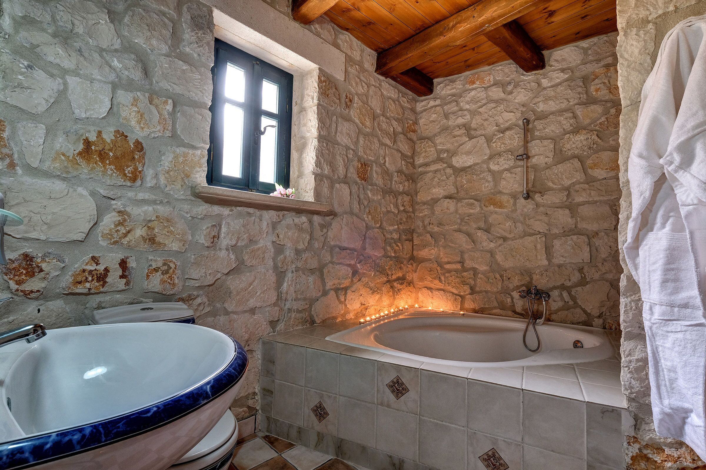 A stone-walled bathroom with a large bathtub, a window, and a white towel hanging on the wall.