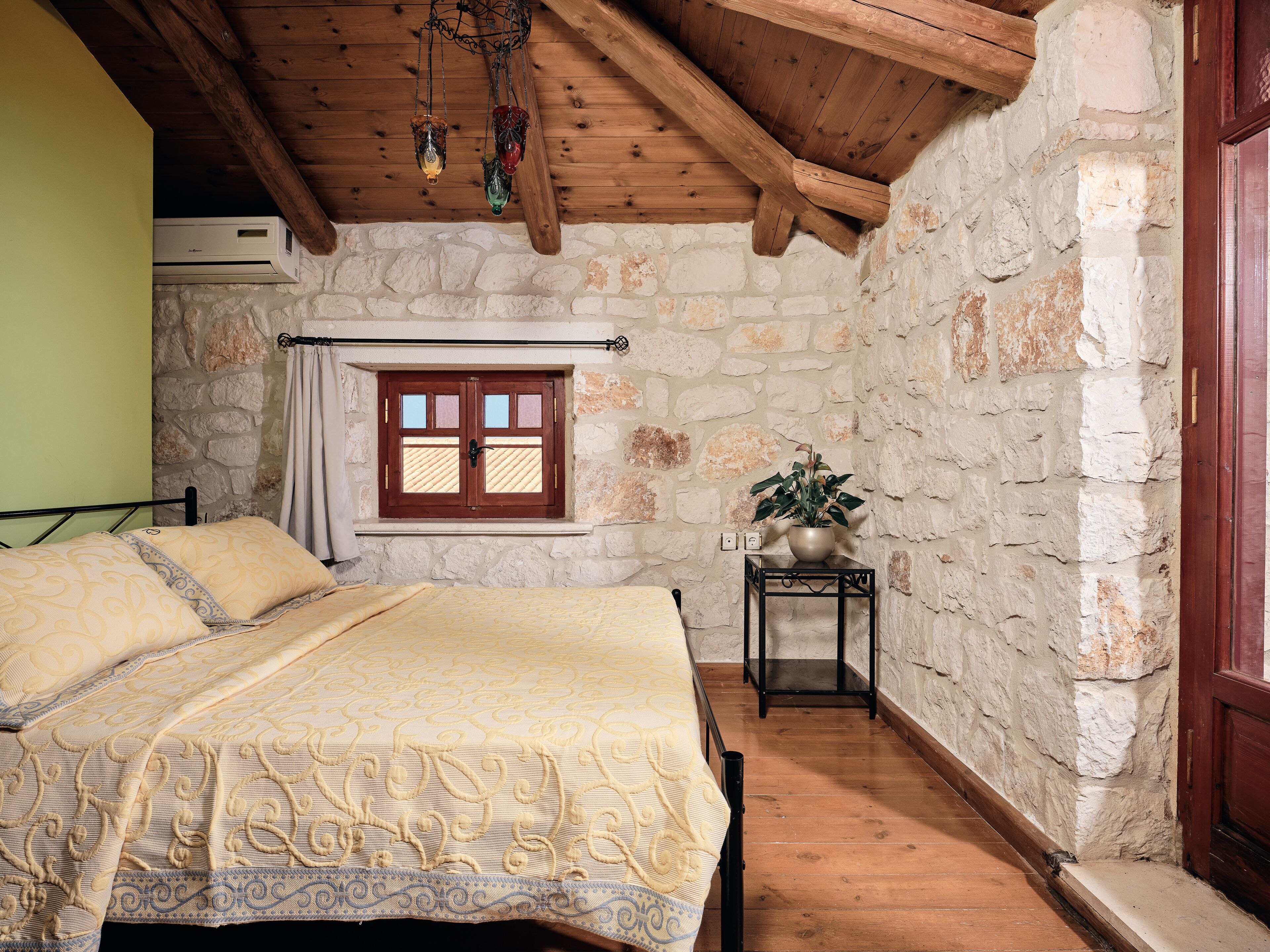 Ein Schlafzimmer mit Steinwand, Holzbalkendecke, einem Bett mit gemusterten Textilien, einem Fenster mit Vorhang und einem kleinen Tisch mit Pflanze.