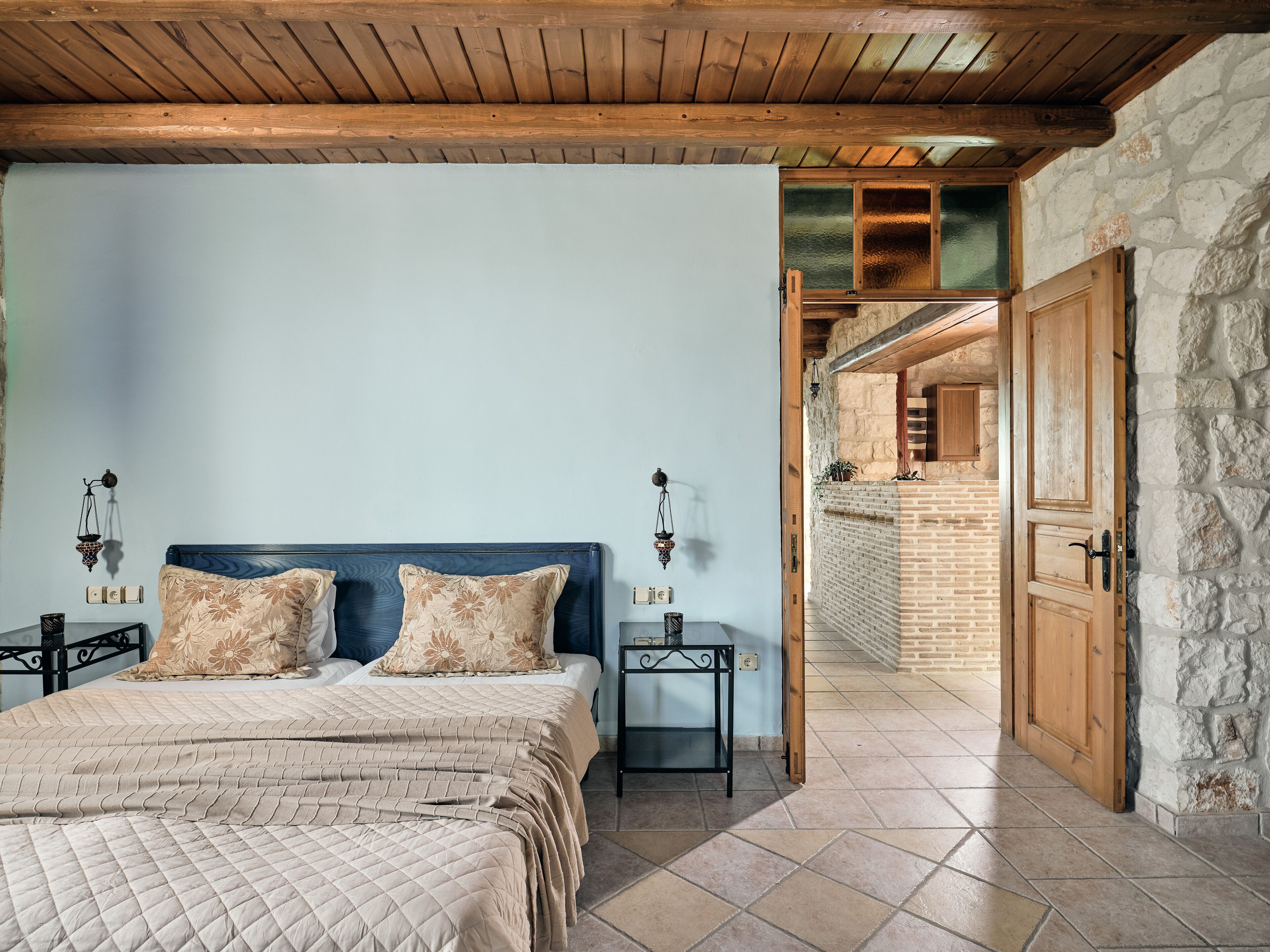 Un dormitorio con techo de madera, una cama con ropa de cama color crema, una mesita de noche y una lámpara de pared.