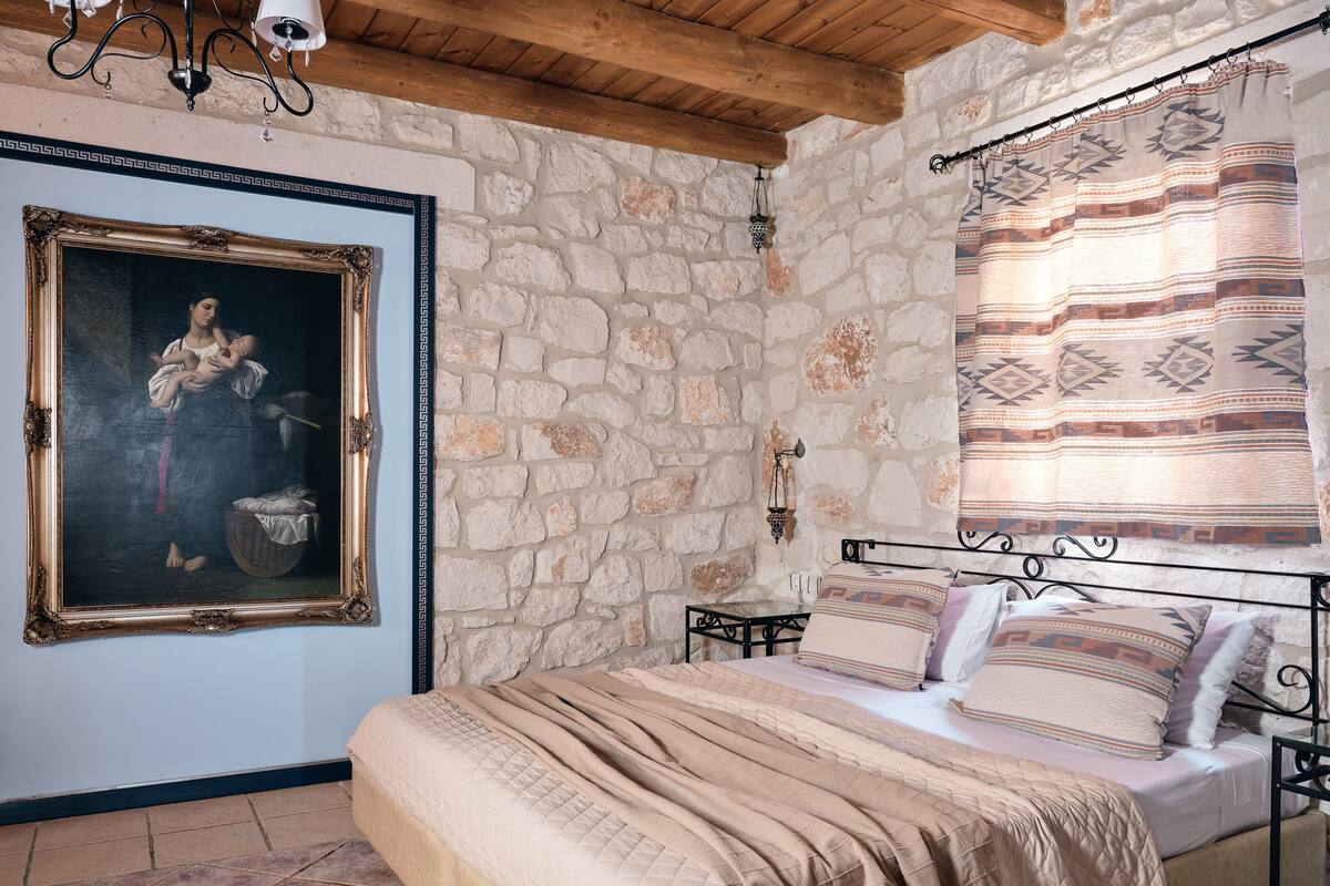 A bedroom with a stone wall, a bed with beige bedding, a painting on the wall, and a window with patterned curtains.