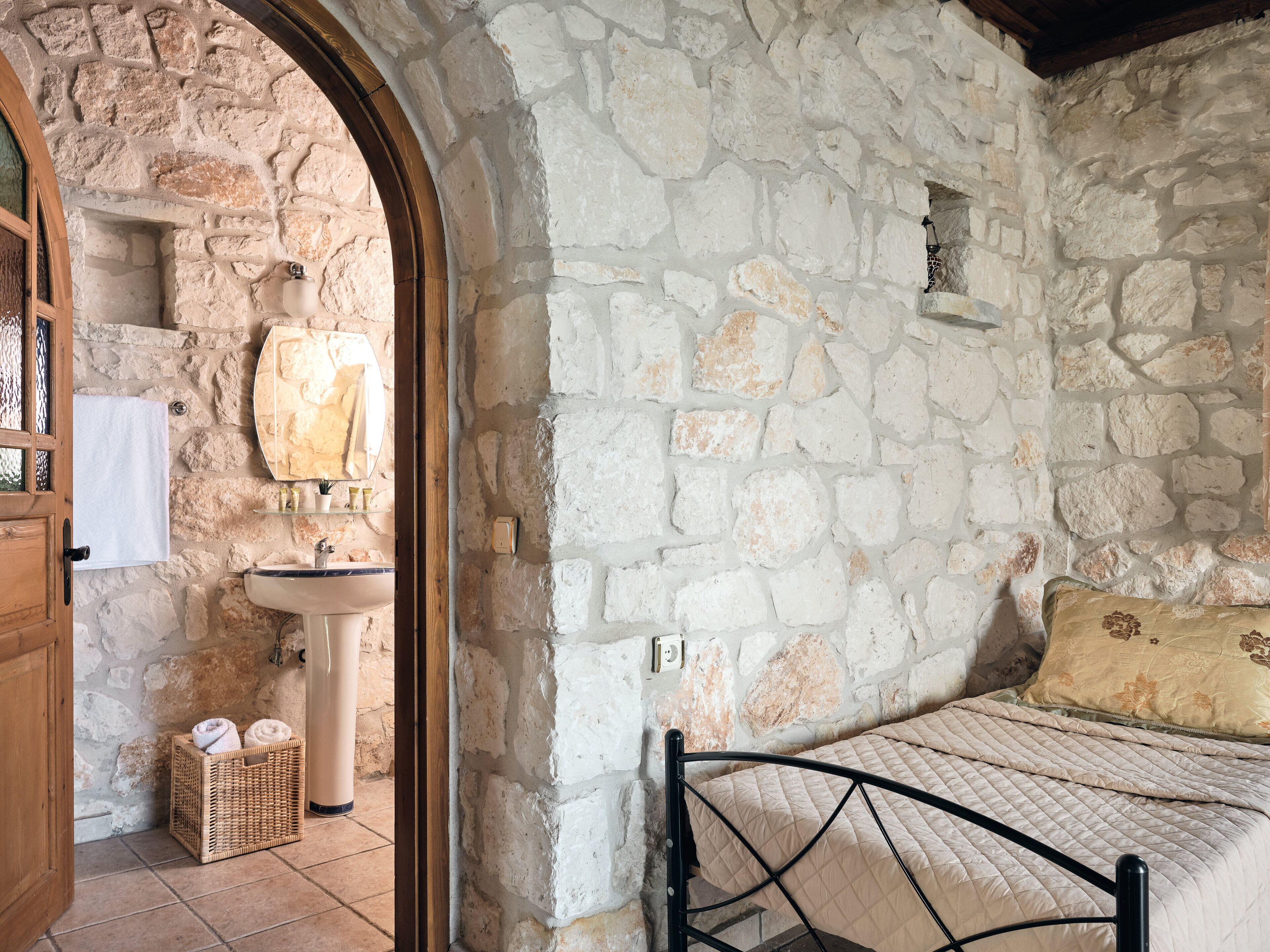 Quarto com paredes de pedra, uma cama, porta de madeira, pia e um cesto de toalhas.