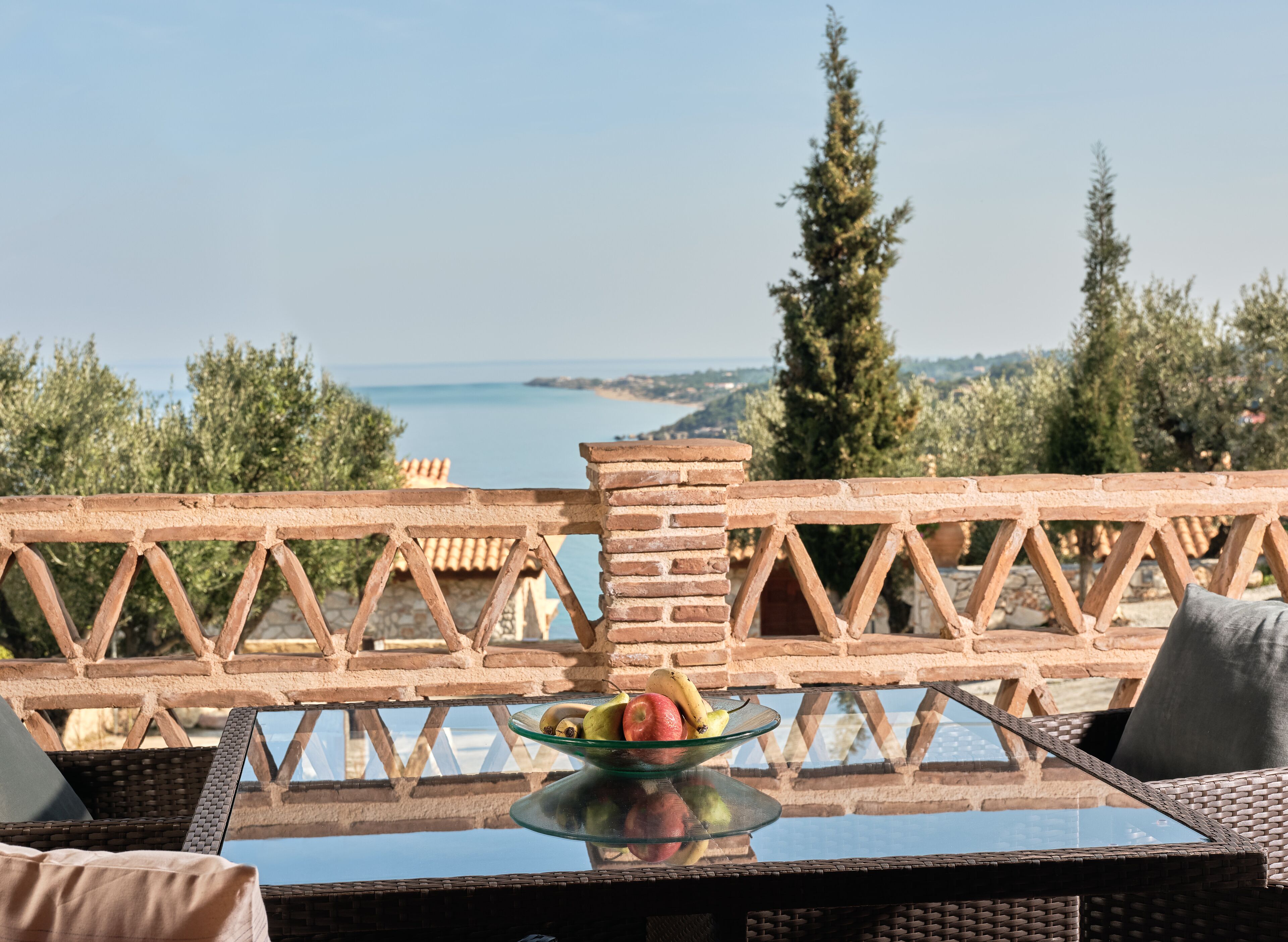 Una mesa de cristal con un frutero, sillas de mimbre y vistas al mar y a los árboles.