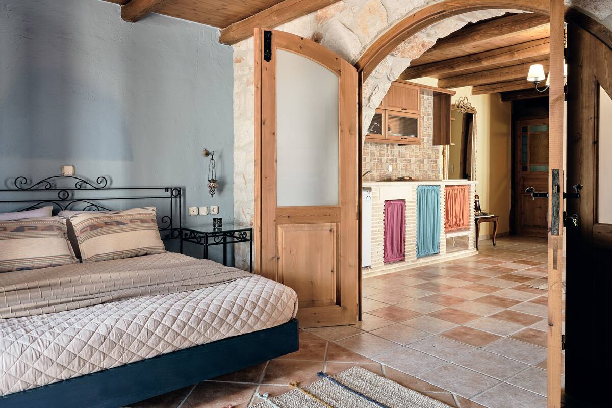 A bedroom with a bed, a nightstand, a wall clock, and a wooden door leading to a kitchen.