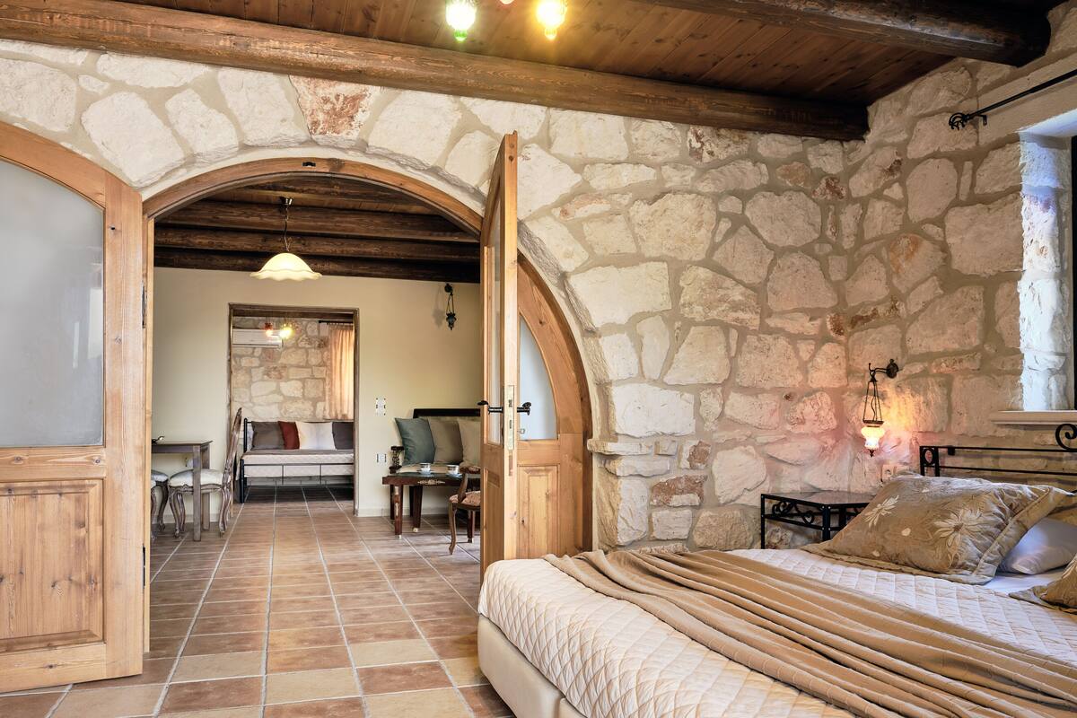 A bedroom with a stone wall, wooden beams, a bed, a table, and chairs.