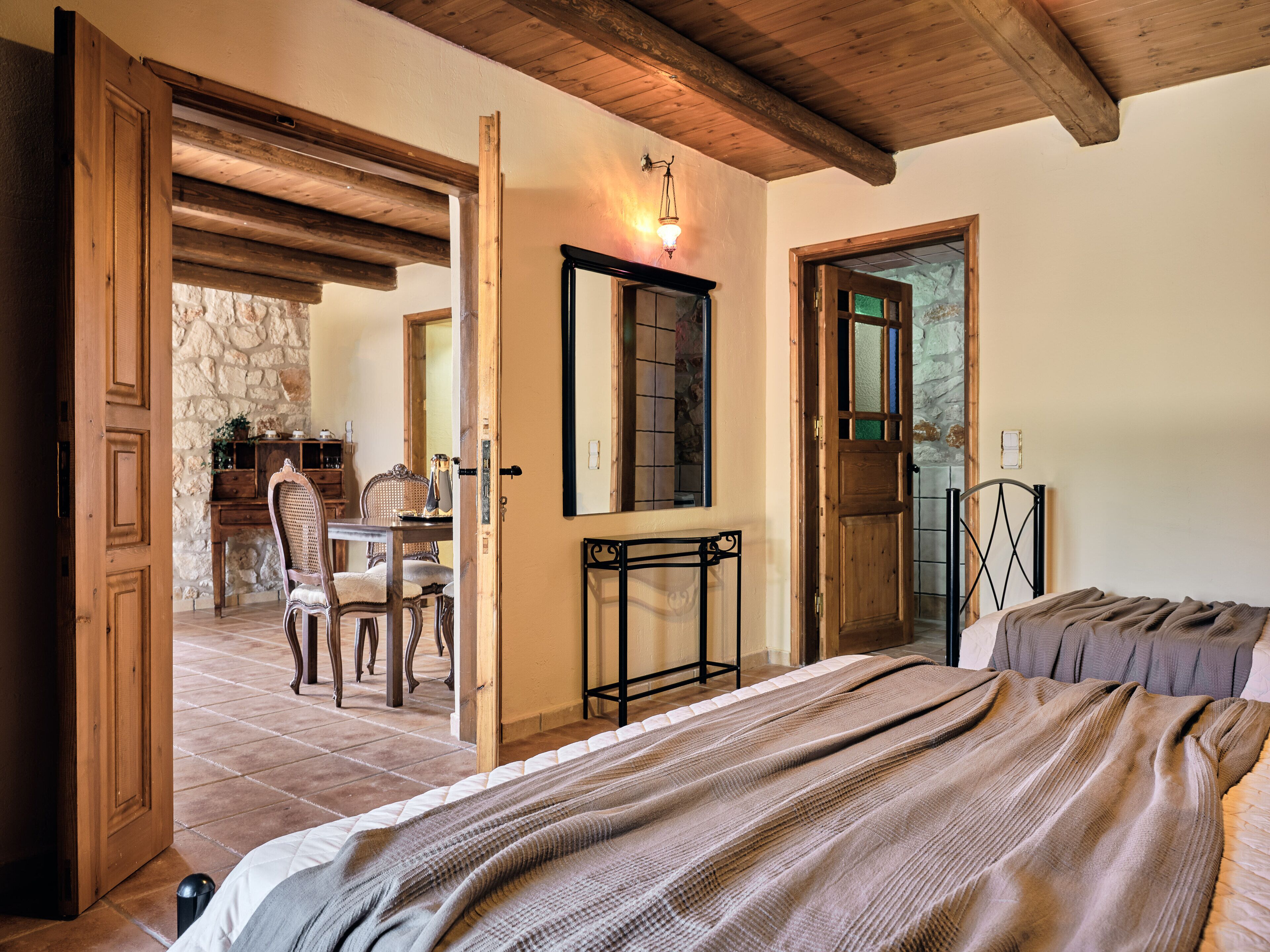 A room with a bed, a wooden door, a stone wall, a dining table with chairs, and a mirror on the wall.