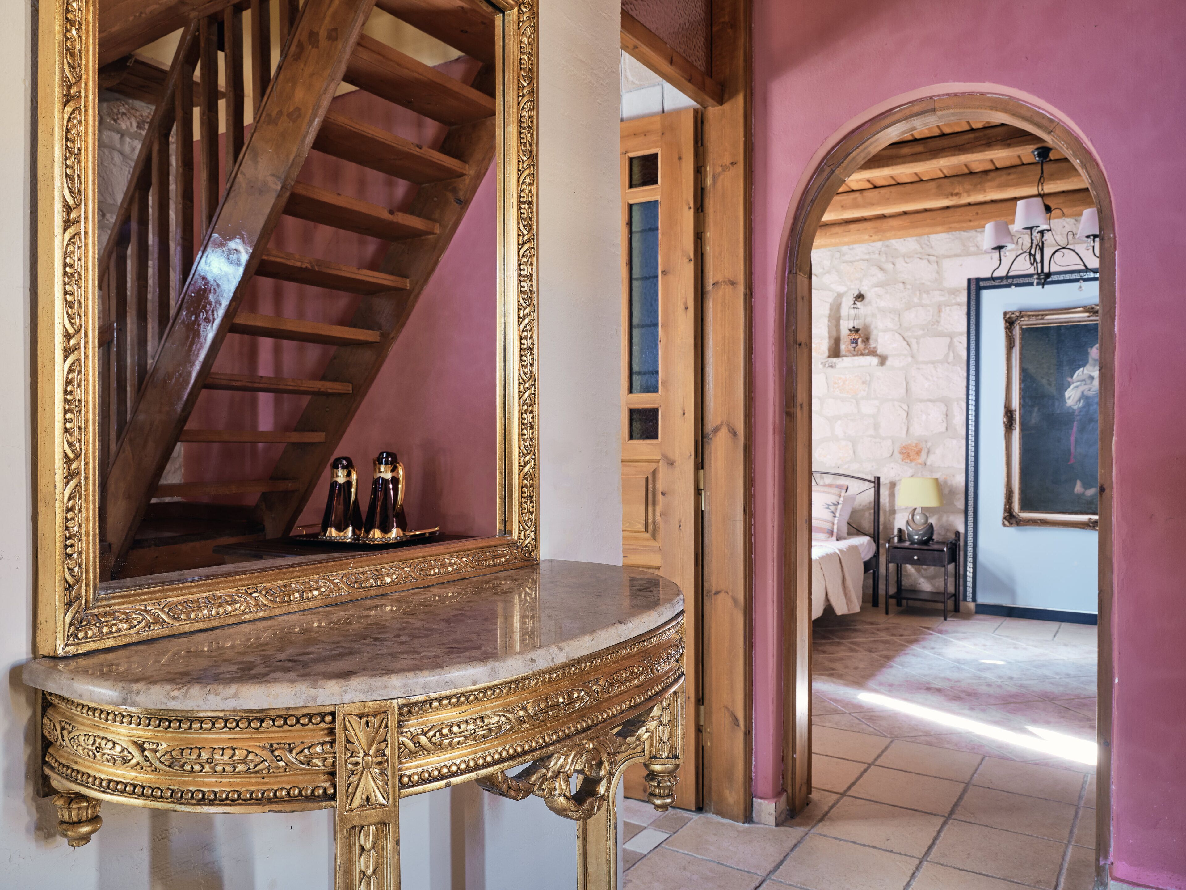 A grand staircase with a wooden finish, a marble pedestal table with intricate gold detailing, and a stone wall in the background.