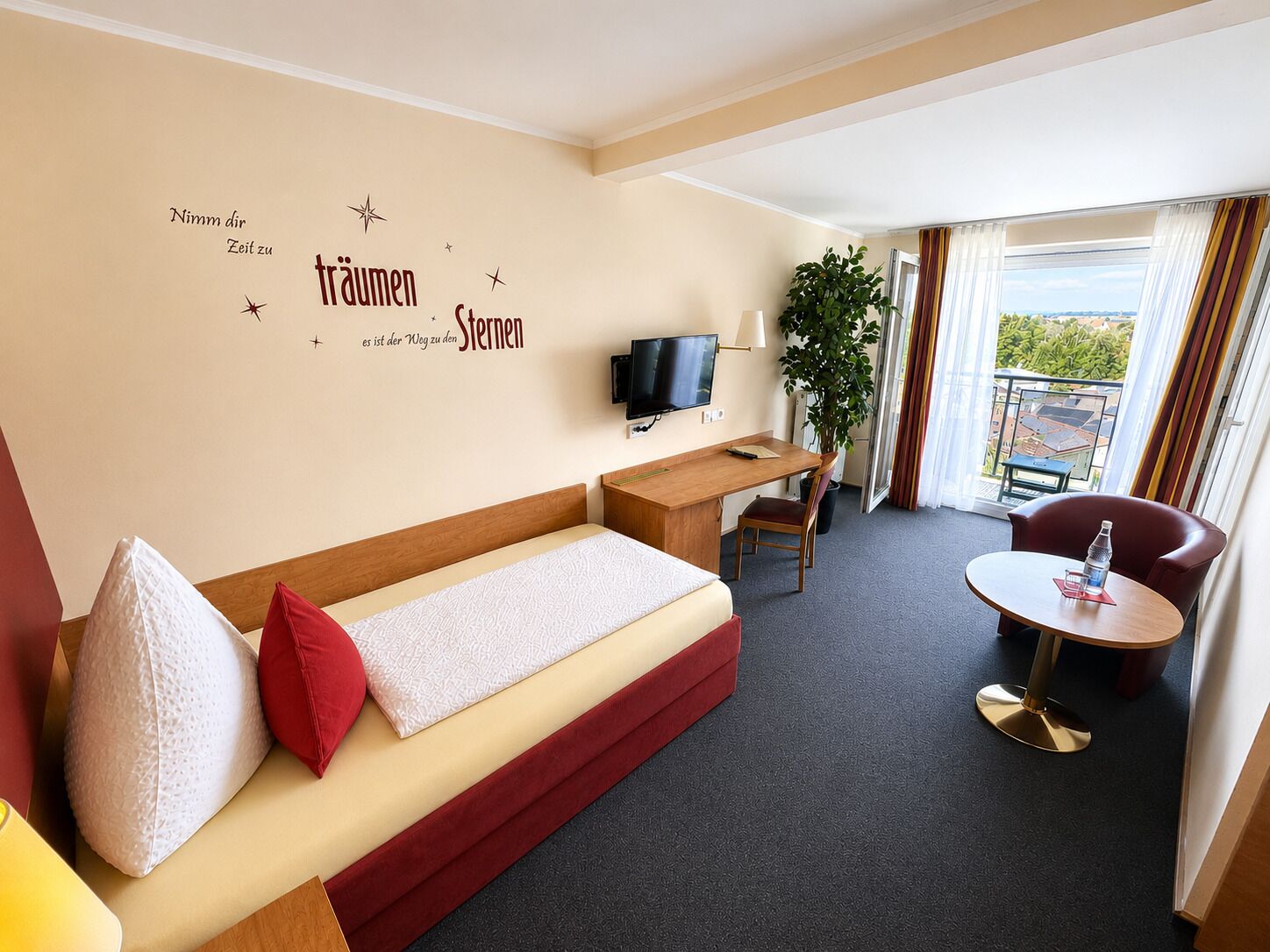 A hotel room with a sofa, a desk, a TV, a chair, a small table, and a balcony with curtains.