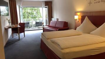 A hotel room with a large bed, a desk, a chair, a red sofa, and a balcony with outdoor furniture.