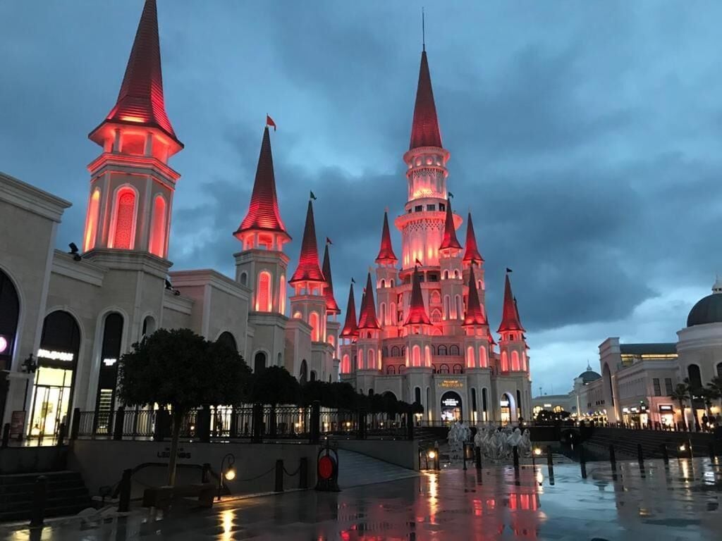 A large, illuminated castle-like building with multiple spires and a central tower, surrounded by a reflective plaza.