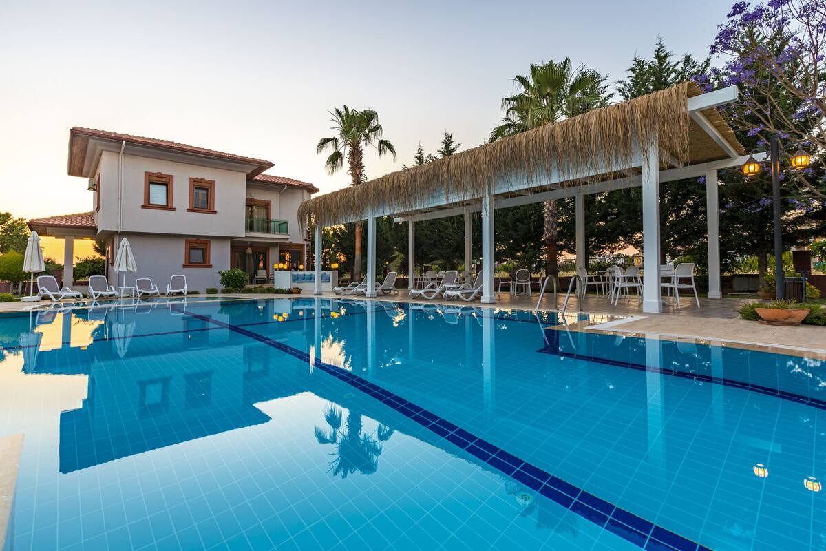 A swimming pool with lounge chairs and a covered area, surrounded by palm trees and a building with a red-tiled roof.