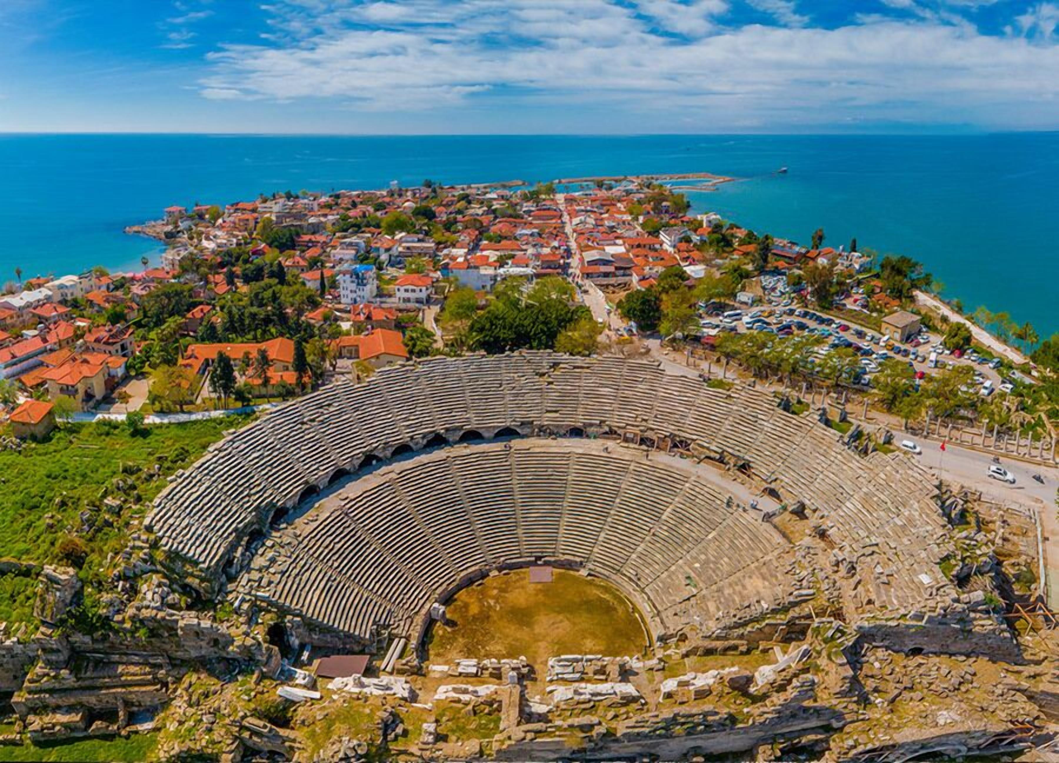 An ancient amphitheater with stone seating and a central stage, surrounded by a coastal town with red-roofed buildings and a clear blue sky.
