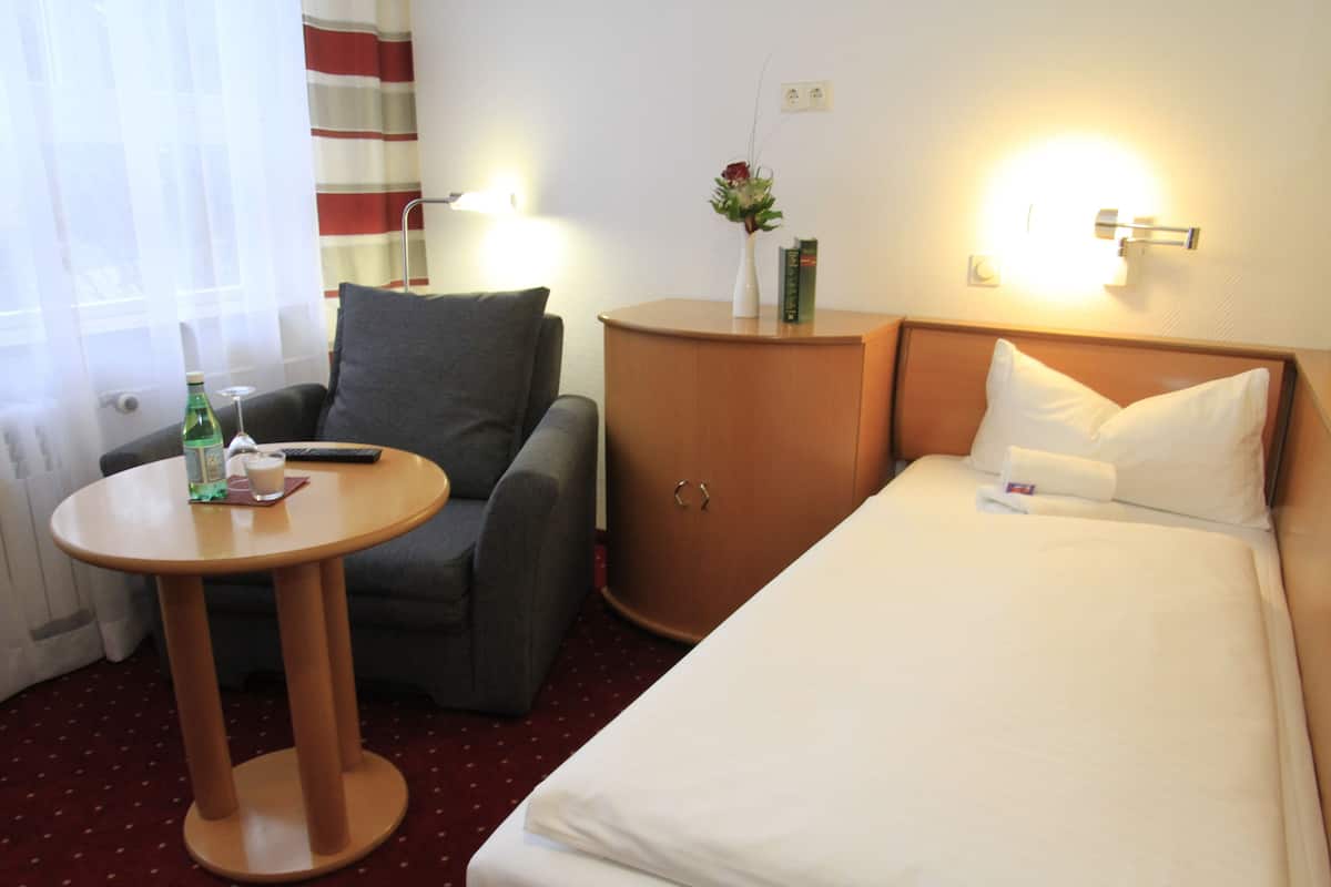 Habitación de hotel con cama, una silla, una mesita, un armario y ventana con cortinas.
