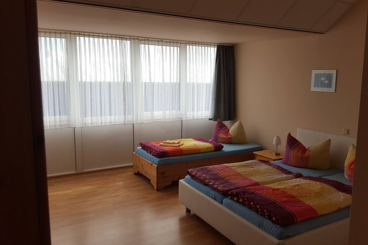 Una habitación con dos camas, una ventana con cortinas translúcidas, un cuadro colgado en la pared y suelo de madera.