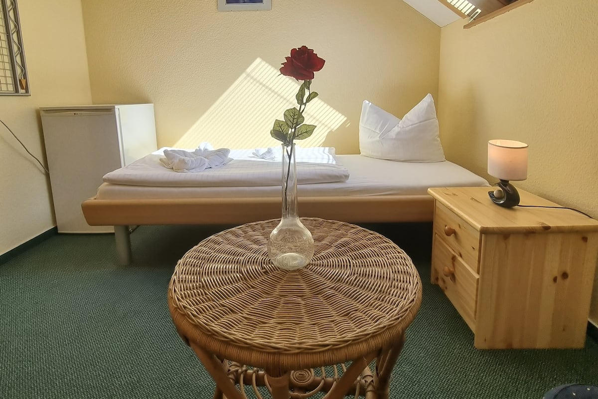 Un dormitorio con cama, una mesita de noche con lámpara, una pequeña mesa redonda de mimbre con un jarrón de una sola rosa roja, y un tragaluz sobre la cama.