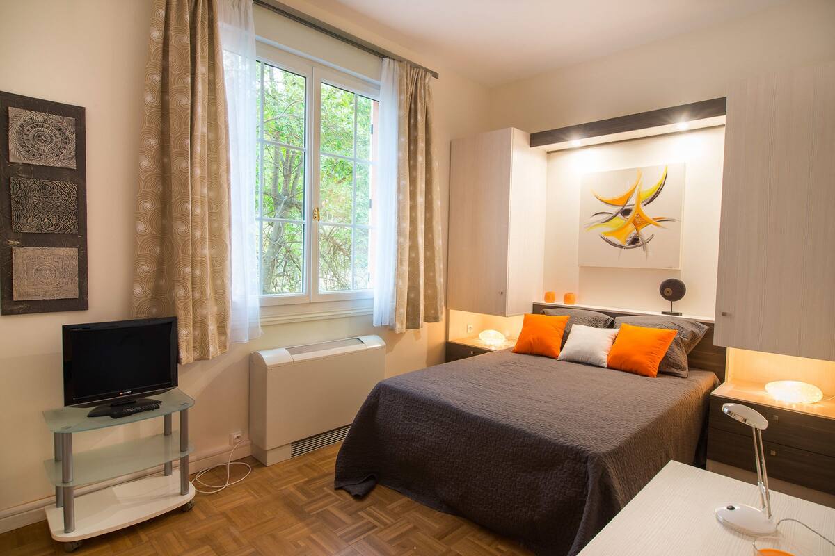 A bedroom with a bed, a television on a glass stand, a window with curtains, a painting on the wall, and a bedside table with a lamp.