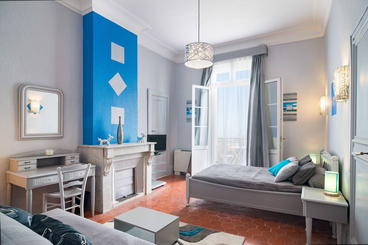 A modern bedroom with a bed, desk, and chair, a large window with curtains, and a blue accent wall.