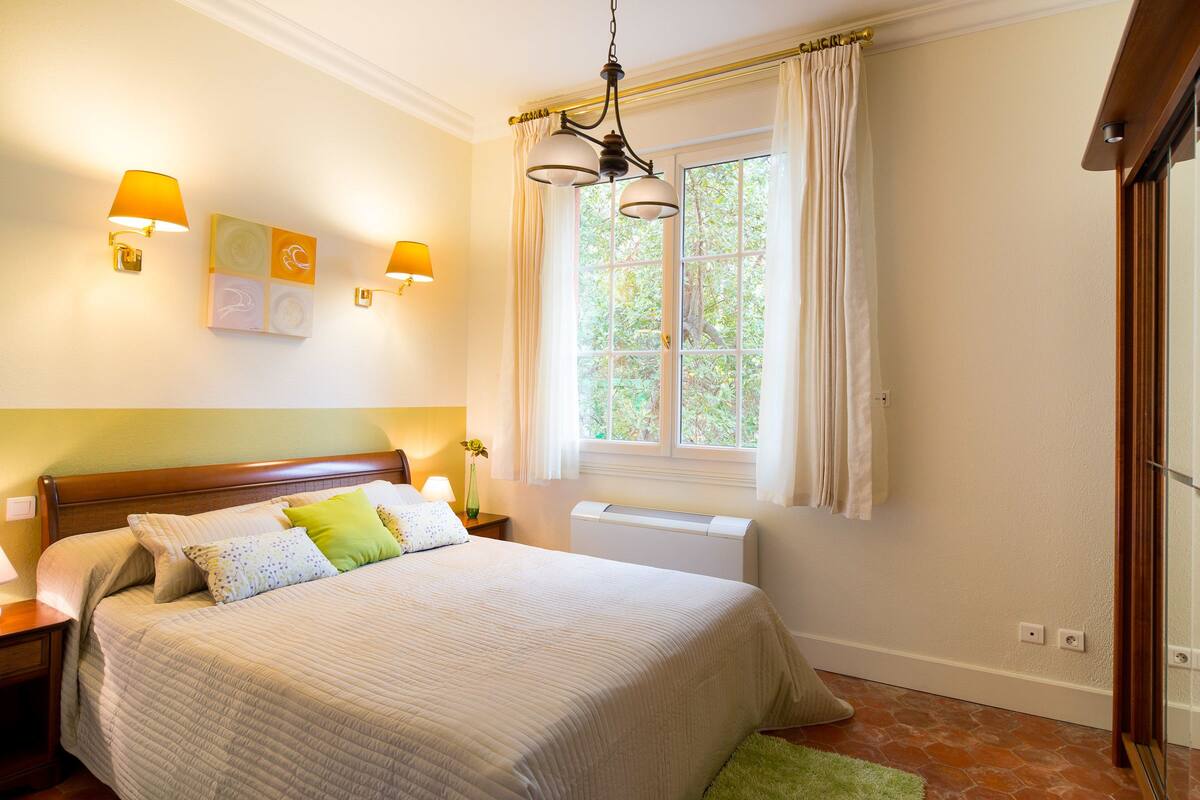 A neatly made bed with pillows, a bedside table with a lamp, a window with curtains, and a radiator.