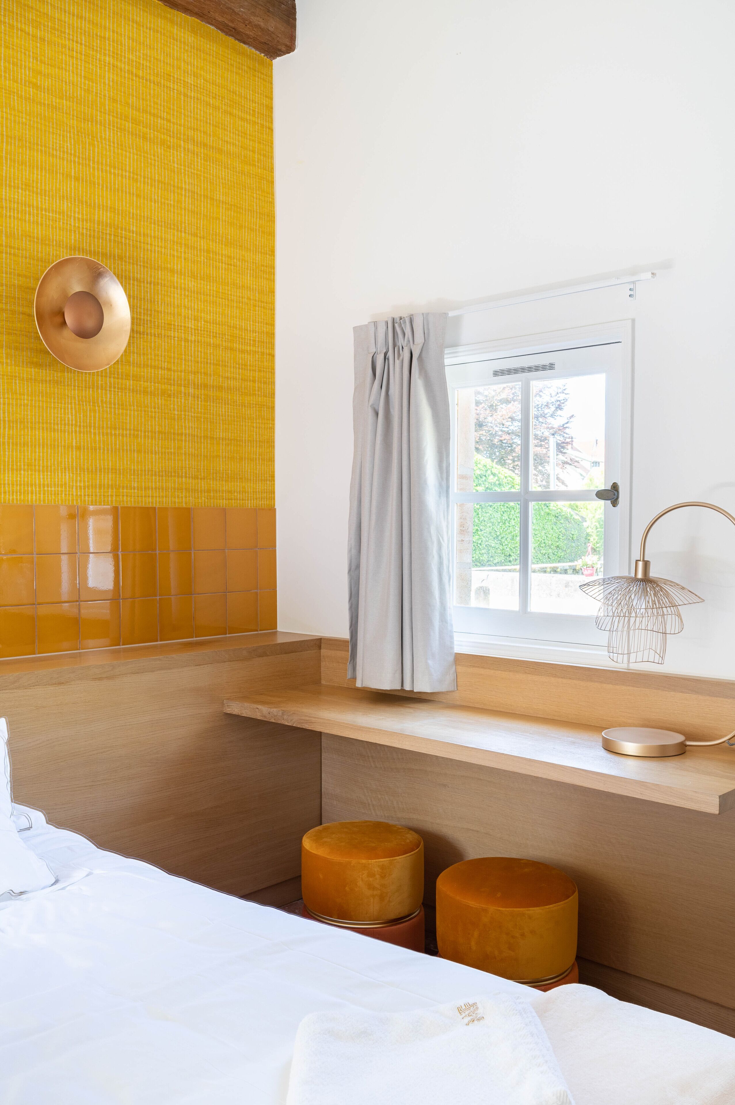 Une salle de bain avec un mur recouvert de carreaux jaunes, une fenêtre avec un rideau, un lavabo et deux tabourets.
