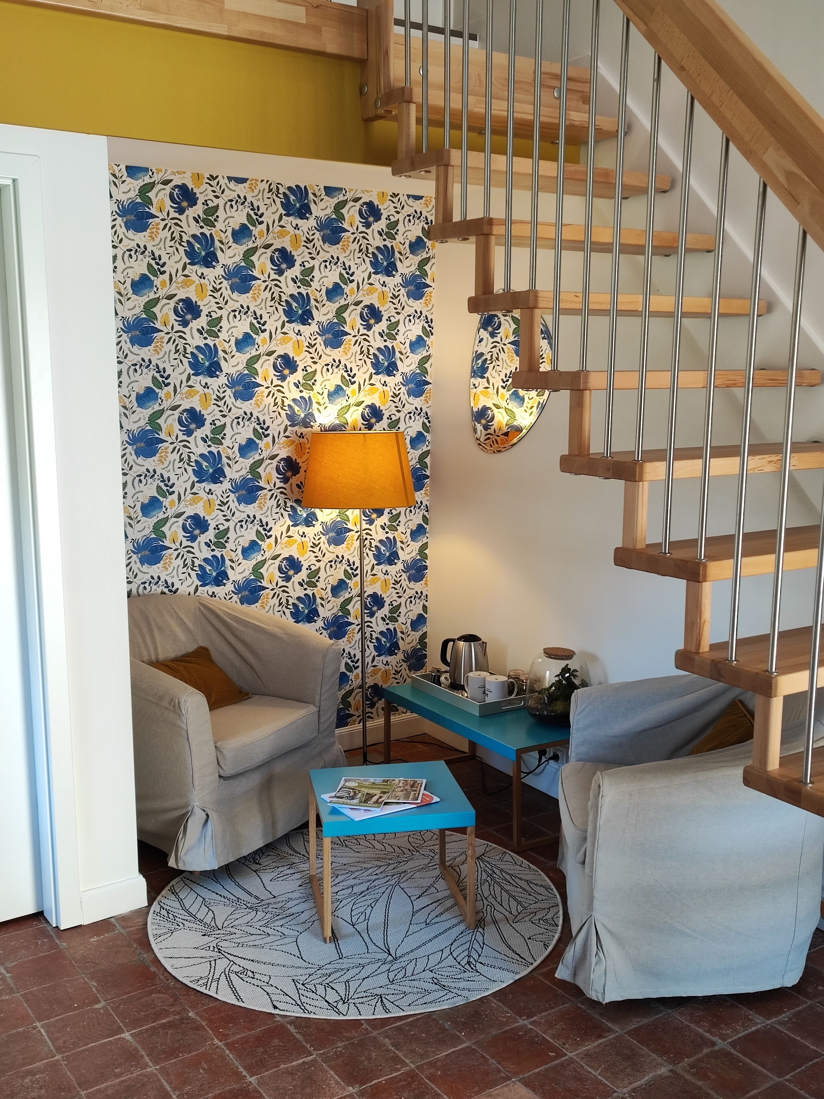 Un salón acogedor con papel tapiz floral, una escalera de madera, una mesa de centro azul y un sofá beige.