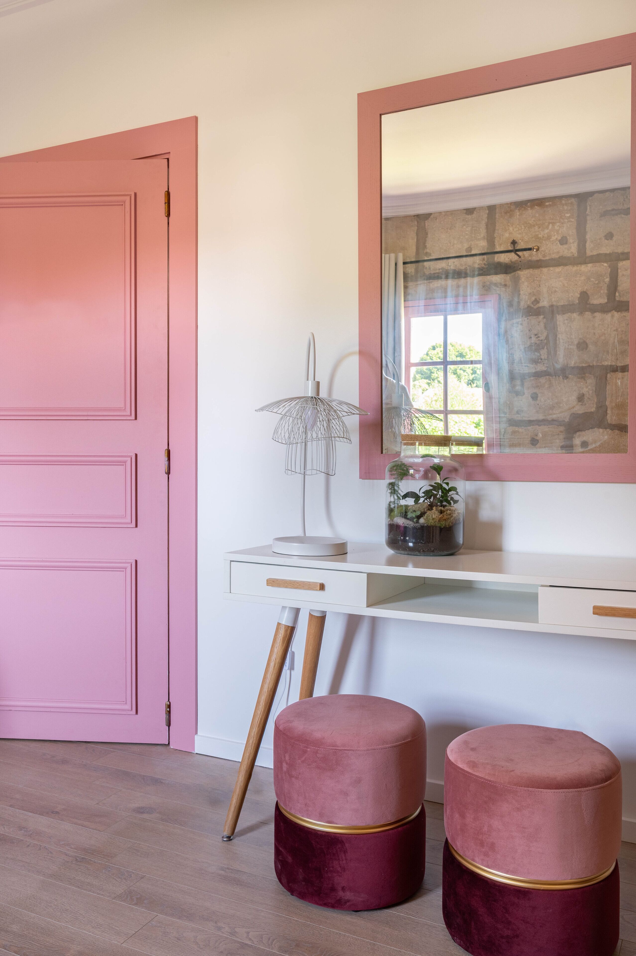 A pink door, a white desk with a mirror, a lamp, and two red velvet ottomans.